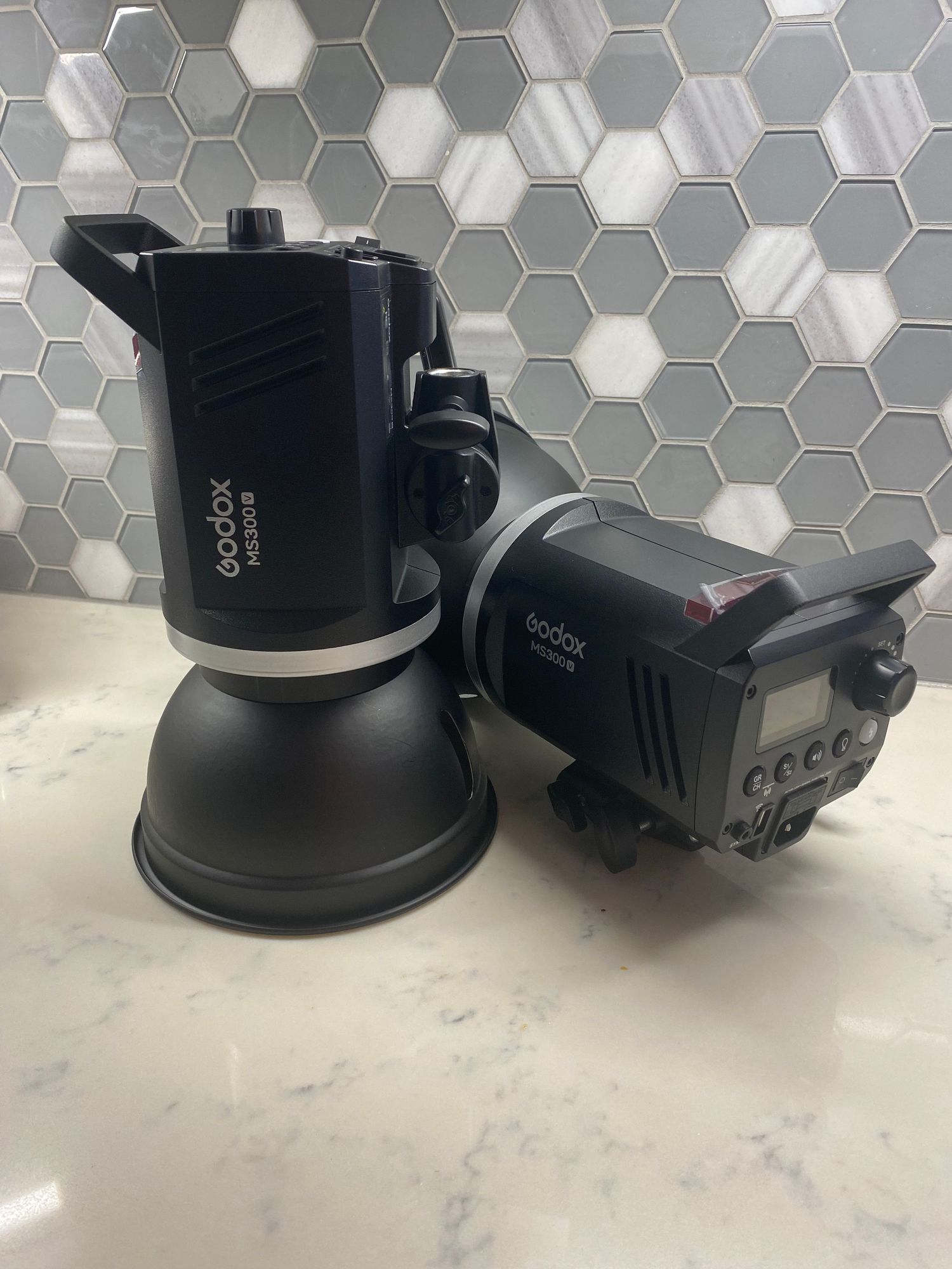 Two black photography lighting flashes displayed against a gray hexagon tile backsplash. Godox MS300V Studio Strobe