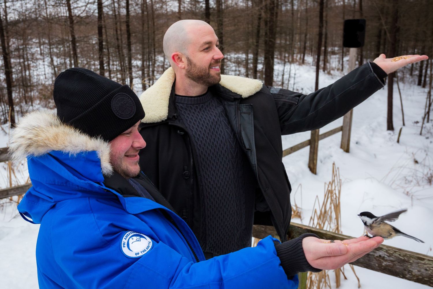 Two men dressed in winter jackets feeding birds in a snowy forest, one holding out their hand as a bird lands on it.