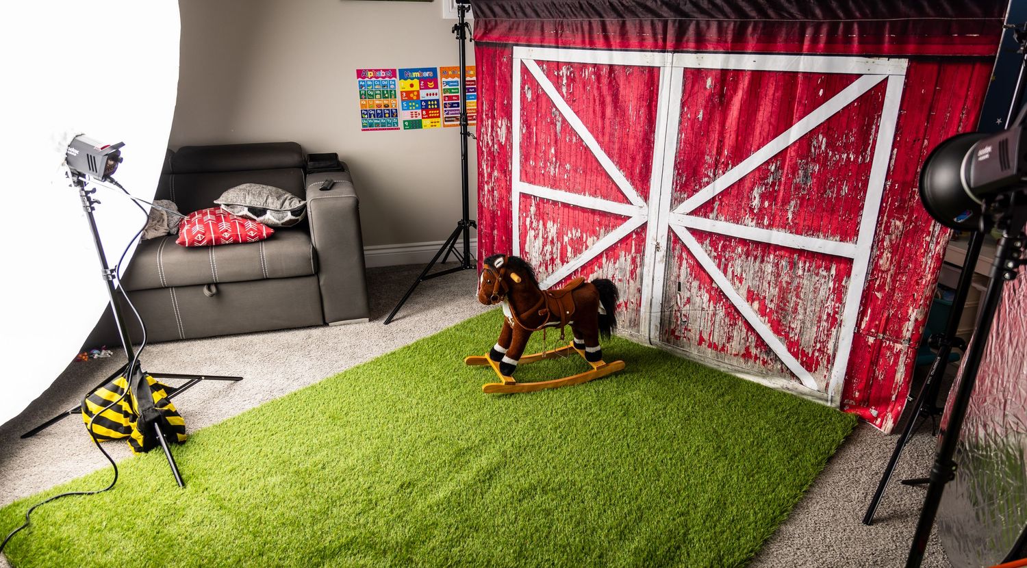 Studio photography setup with red barn backdrop, artificial grass, and rocking horse props.