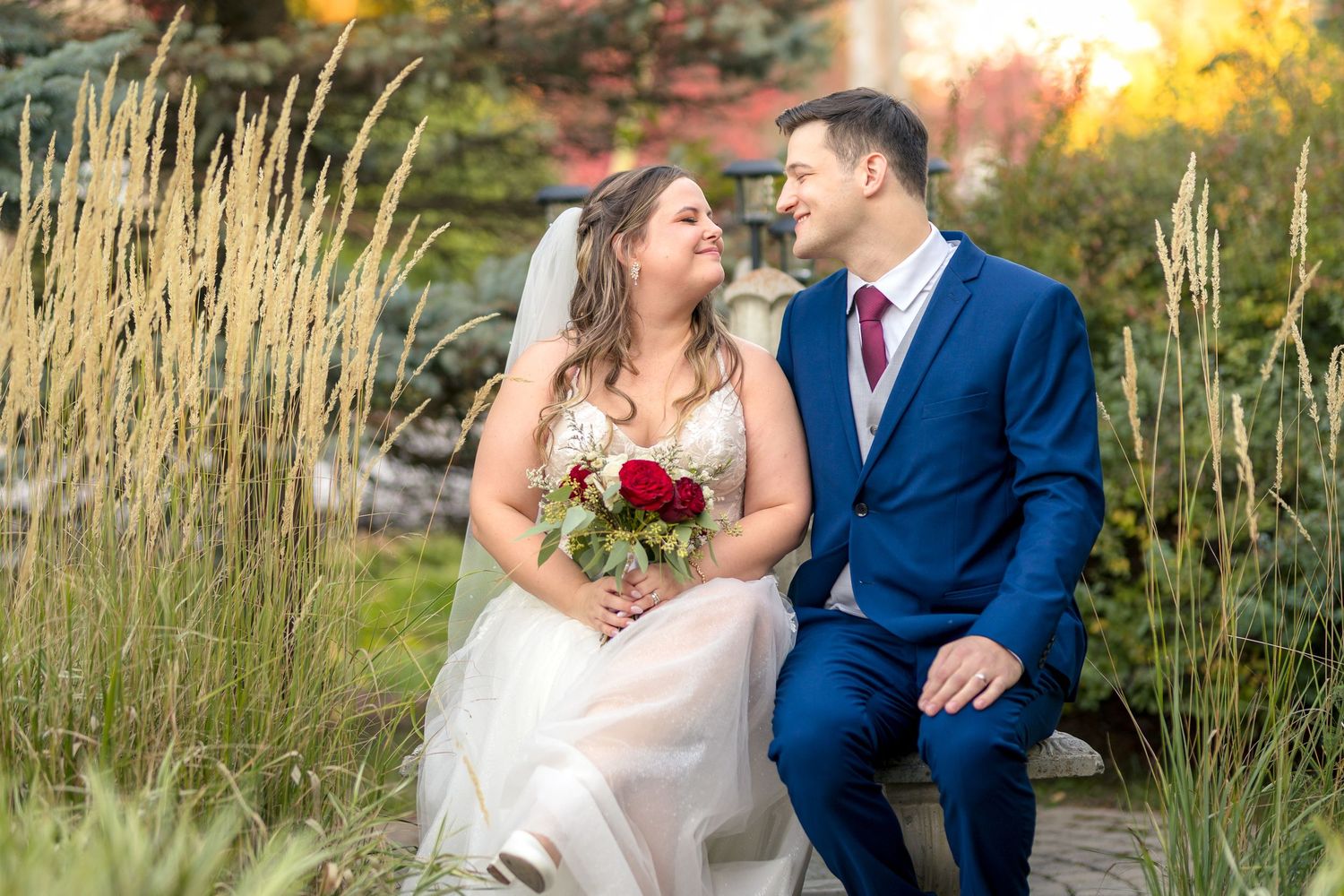 A couple in wedding attire embraces in a garden setting with golden sunlight and tall grass in the background At the school house Munster