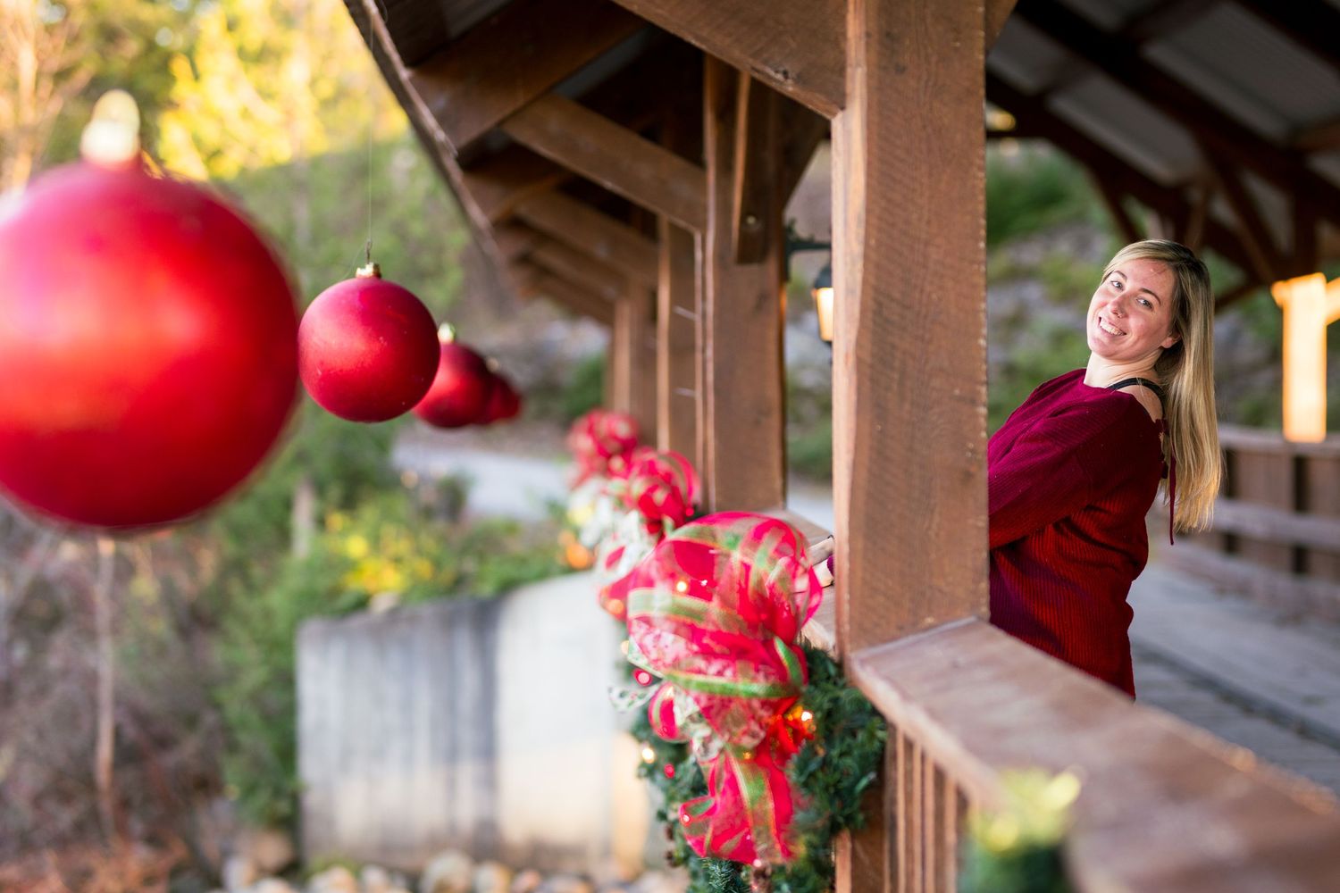Smiling woman on a holiday-decorated wooden bridge with red ornaments and garlands.