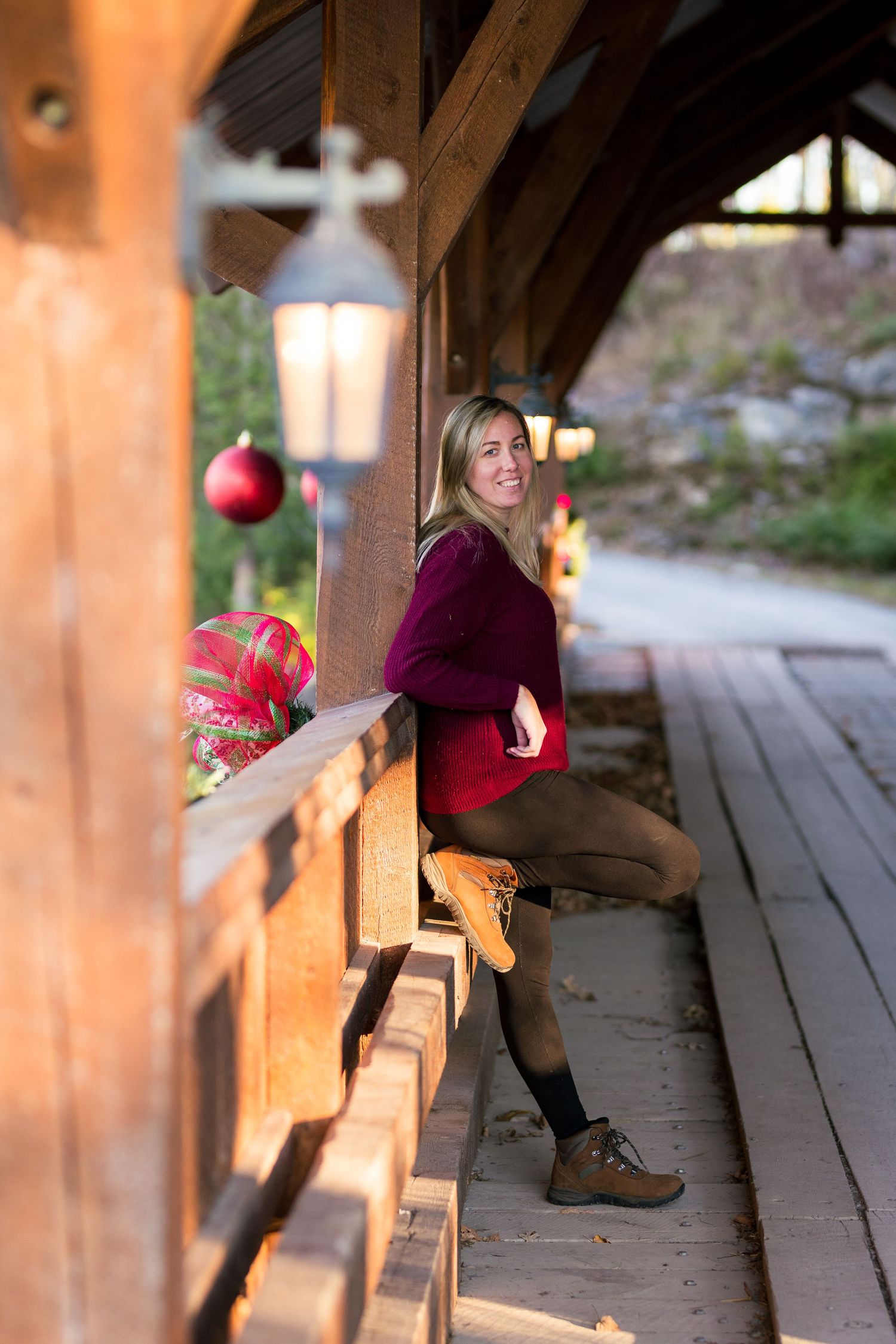 A person in a burgundy sweater and boots sits on a rustic wooden porch decorated with holiday ornaments and lanterns.