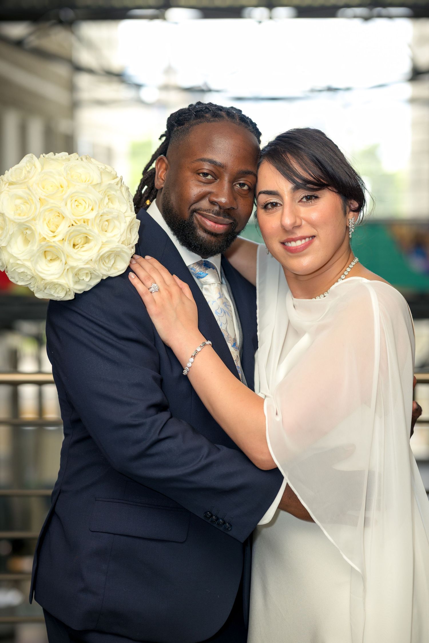 Interracial couple in wedding attire smiling, groom in navy suit, bride in white dress holding bouquet of white roses. Ottawa Wedding Photo