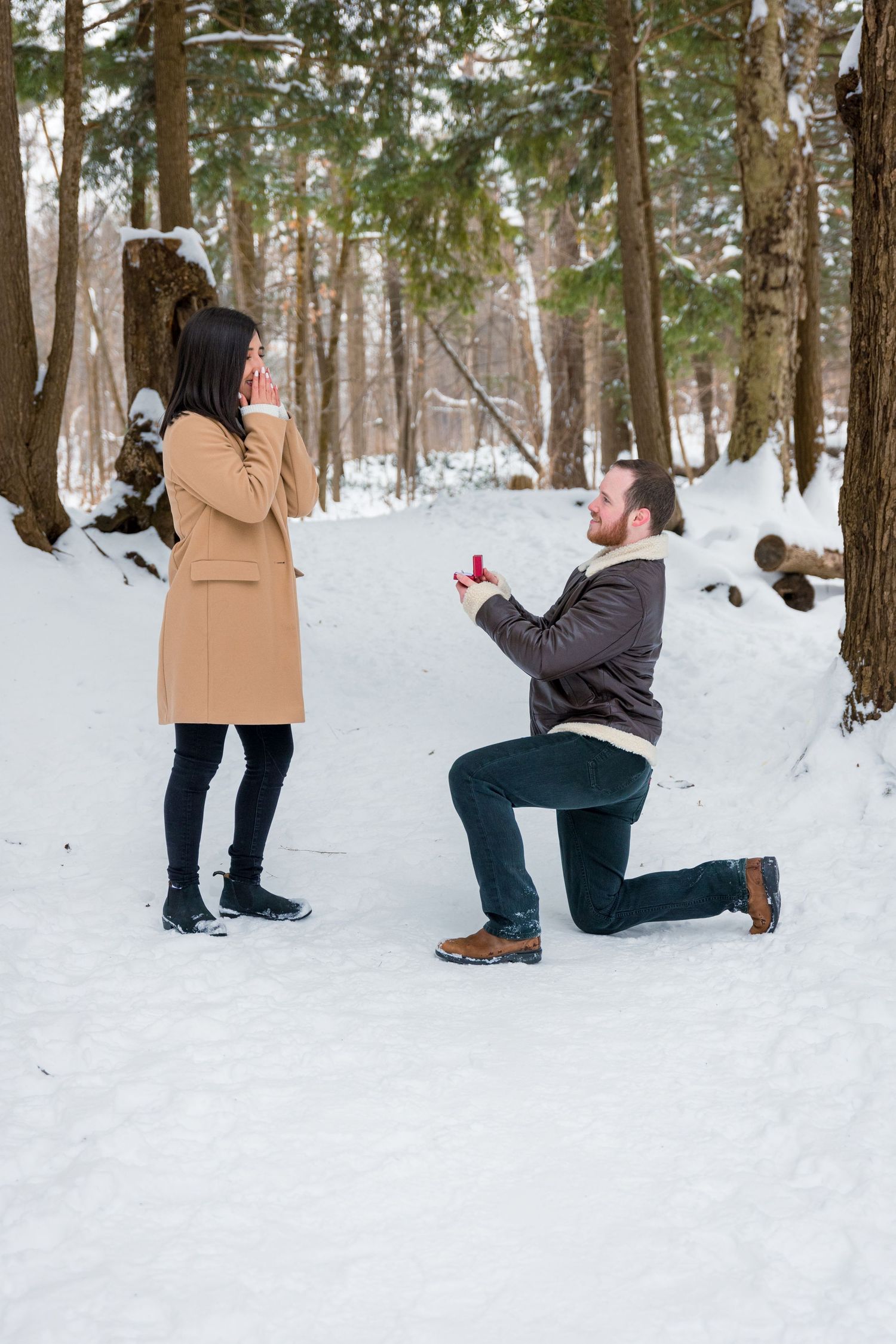 The moment of the proposal: the man down on one knee in the snow, holding out a ring box, as his partner looks on in joyful surprise.