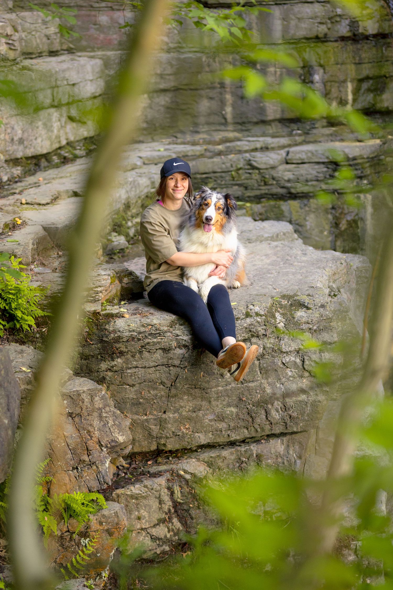 Dog and companion rest on rocky cliff framed by green foliage and waterfall. Ottawa Portrait Photographer