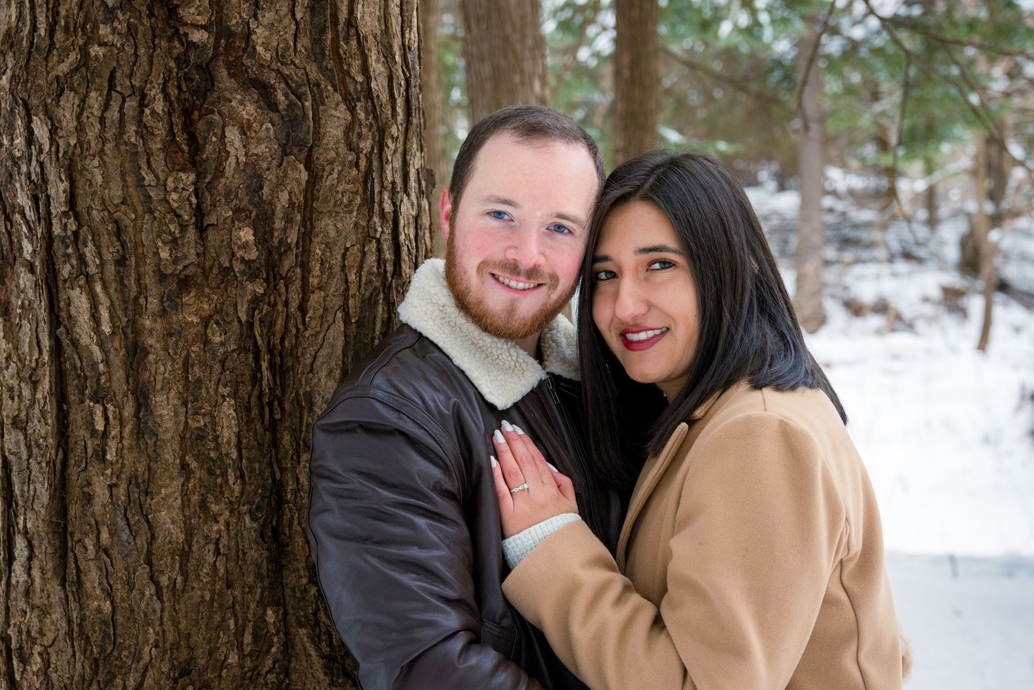 A happy couple poses together by a large tree trunk in a snowy winter forest landscape.