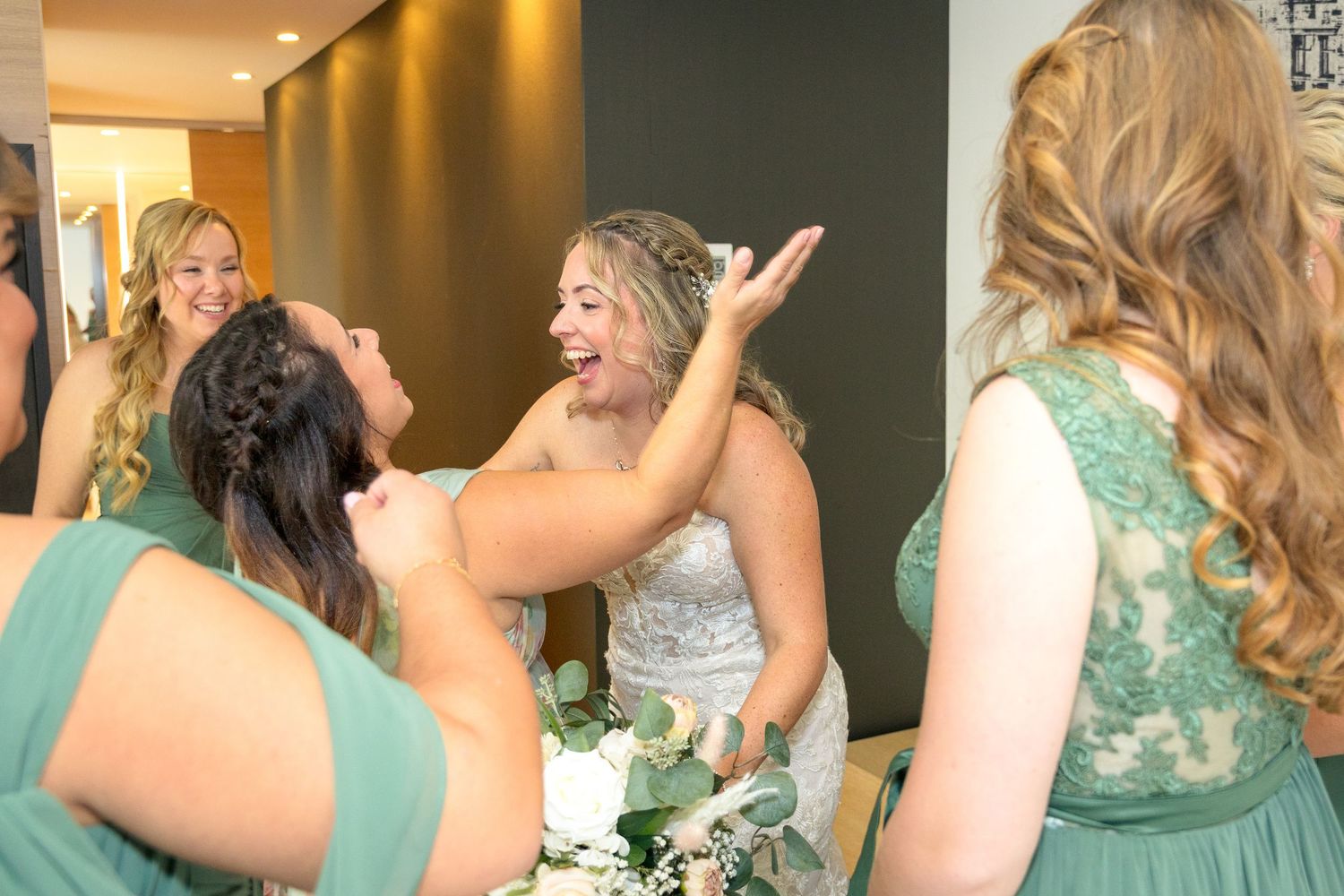 Bridesmaids in mint green dresses gather excitedly around the bride in a warmly lit hallway. Ottawa wedding photographer near me