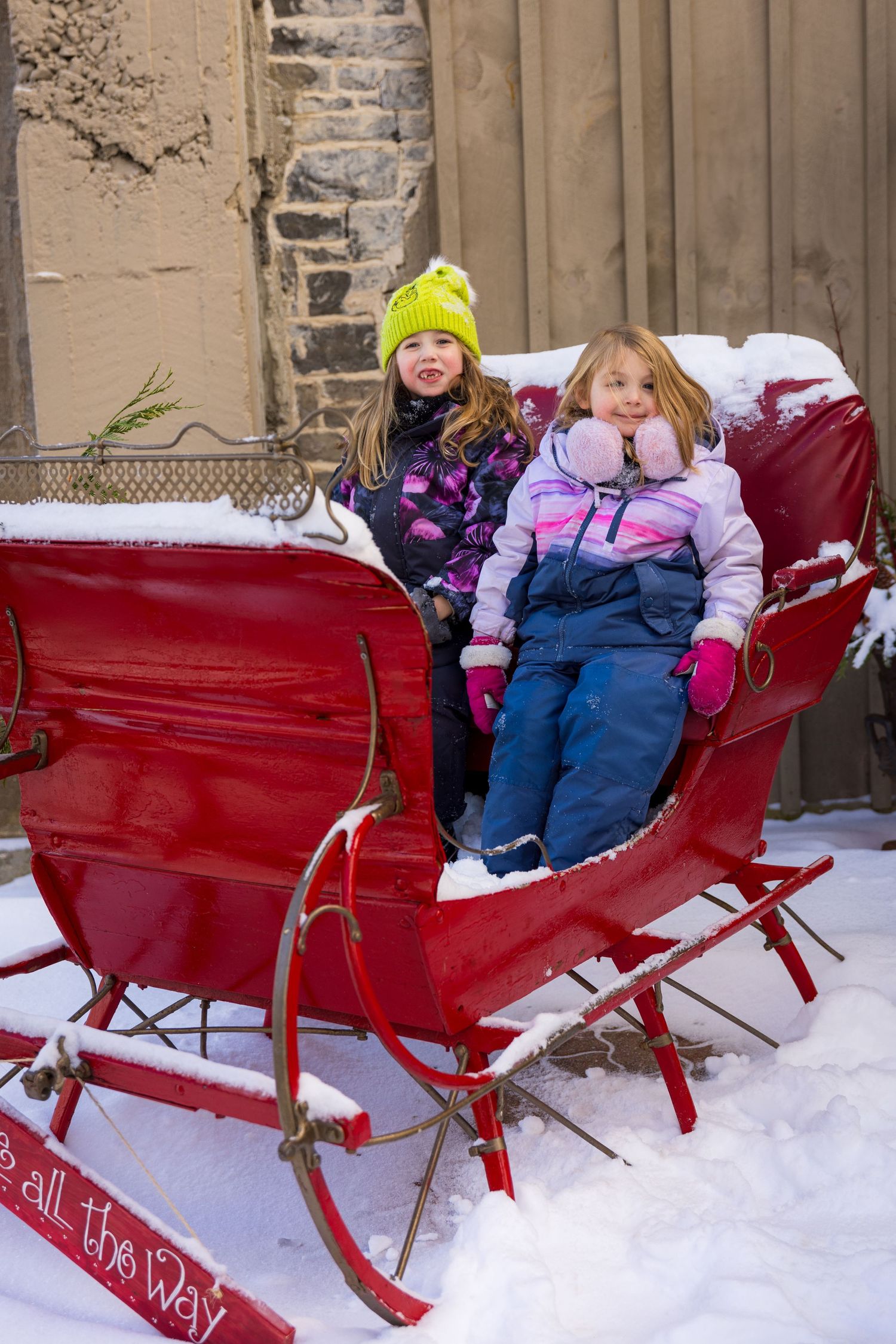 Children bundled up in winter clothing sit in a bright red vintage sleigh in the snow.