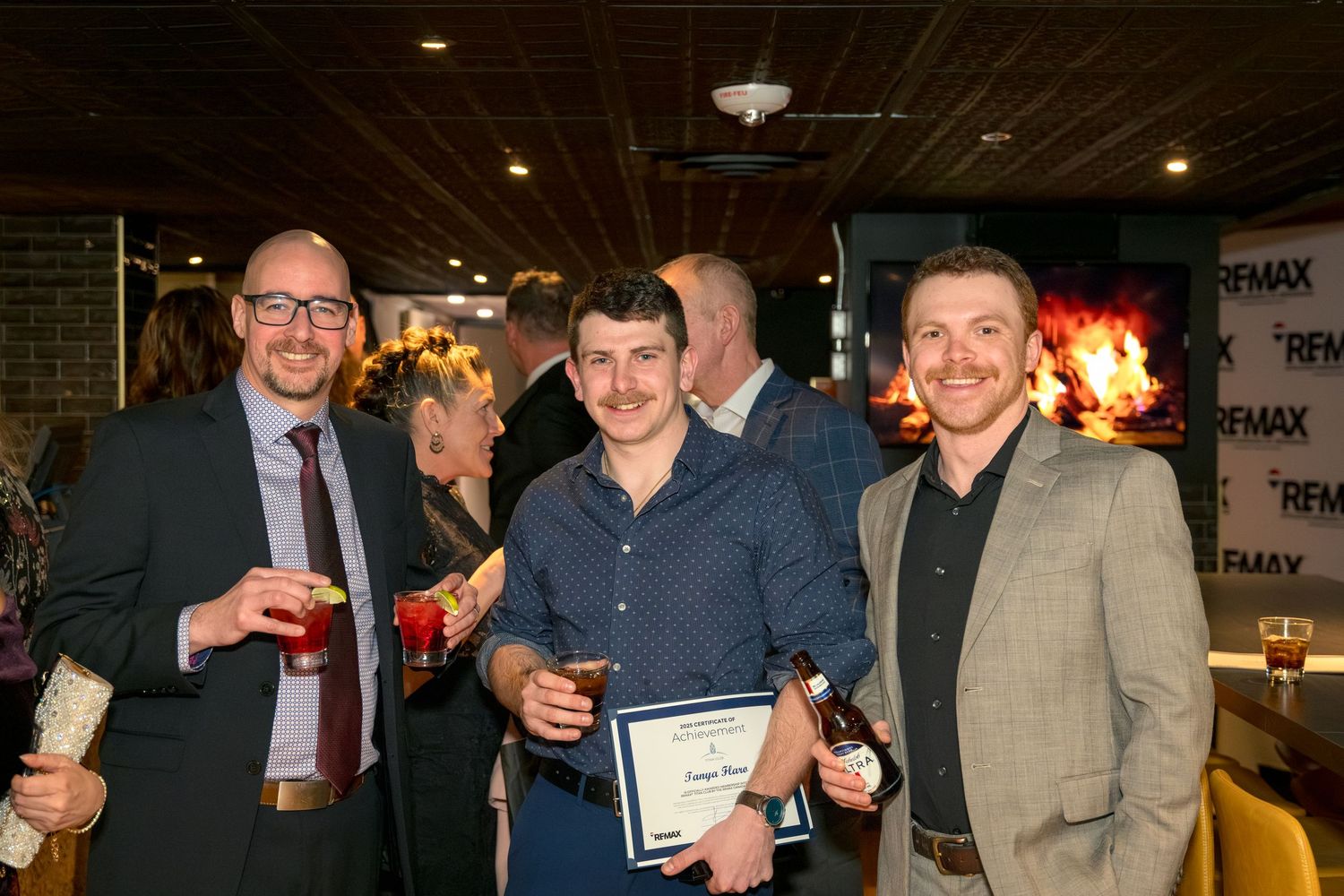 A group of people holding drinks and certificates gather at a dimly lit bar or restaurant for a celebration. Corporate Photographer