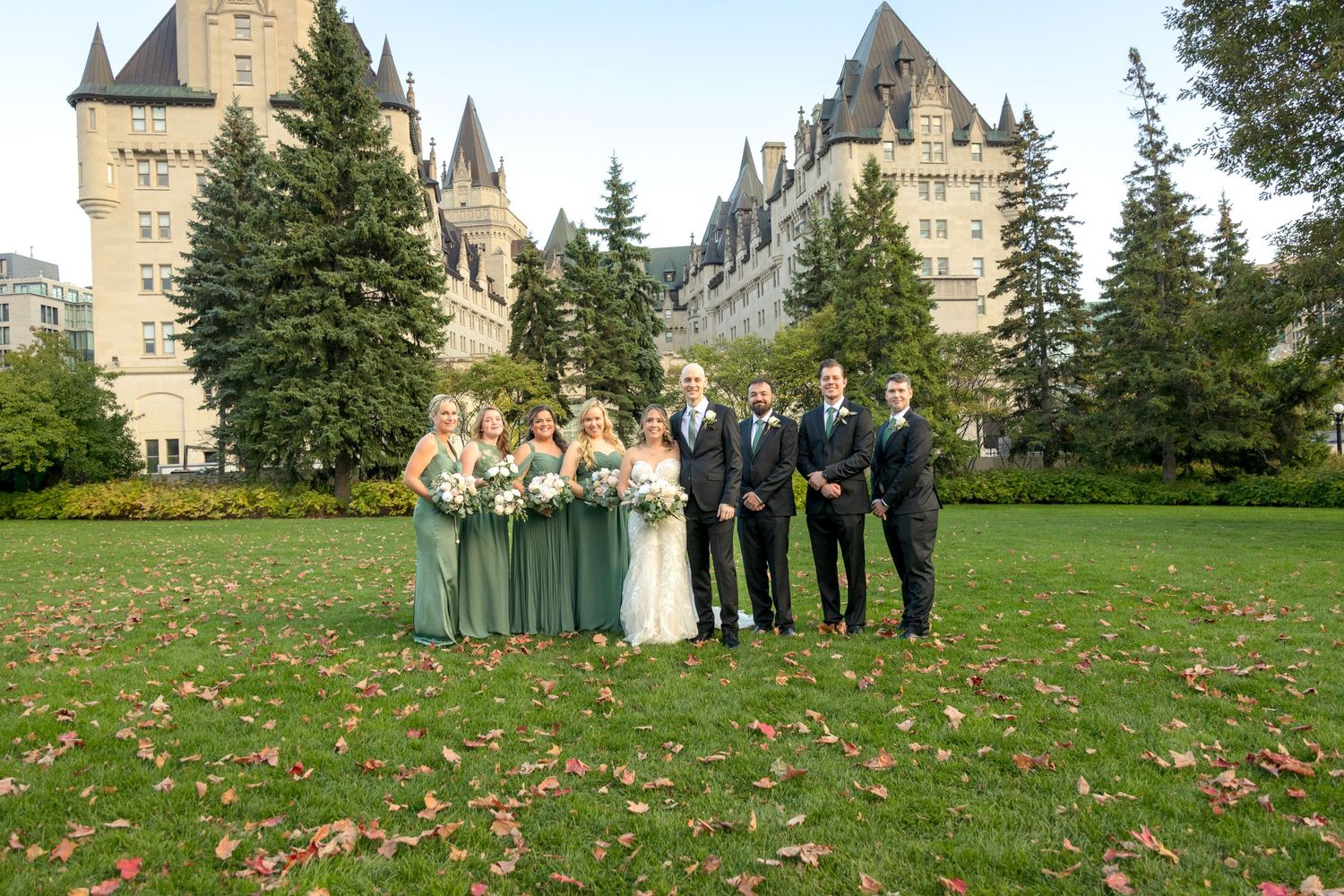 Wedding party poses for photos on a green lawn behind the Fairmont Château Laurier and evergreen trees in the background. Ottawa Wedding