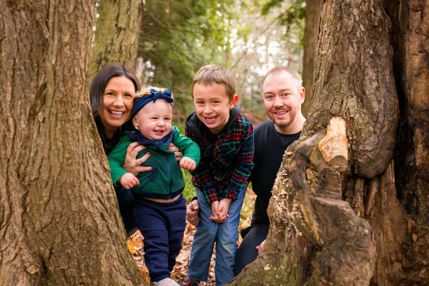 Group poses playfully between large tree trunks in autumn forest setting during outdoor photo session.