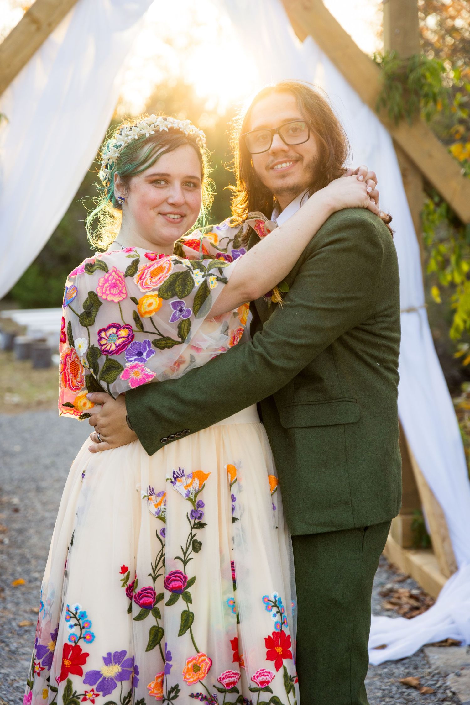 A couple embraces in front of a white fabric backdrop with sunlight behind them, wearing floral and formal attire.