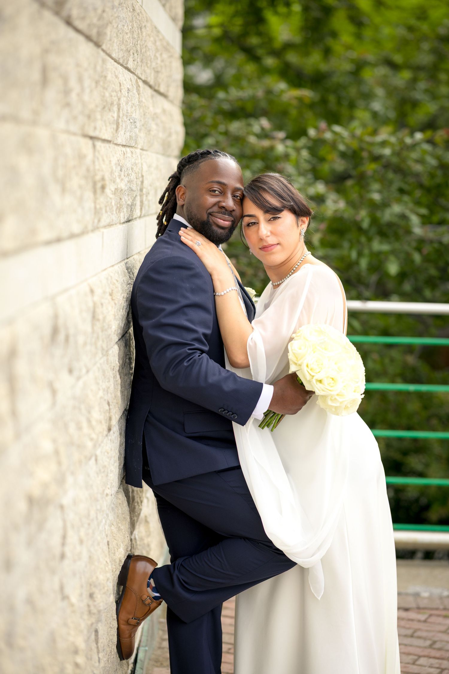 Interracial couple in wedding attire embracing by a stone wall, bride holding white bouquet. Ottawa Wedding Photographer