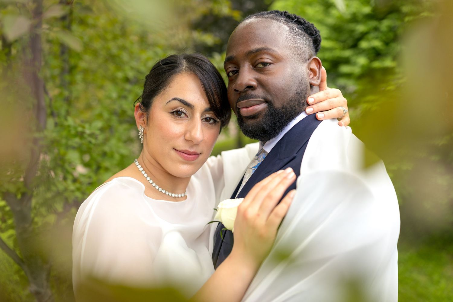 Interracial couple in wedding attire embracing outdoors, bride in white dress with pearls, groom in suit, green foliage background. Ottawa