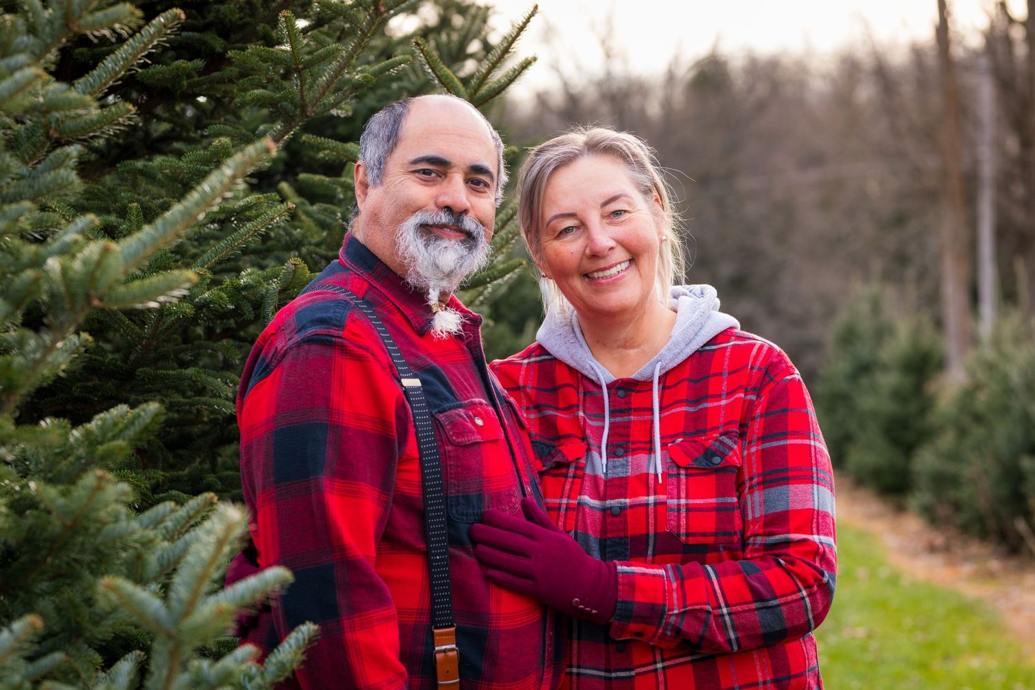 Couple in matching red plaid flannel shirts standing together by evergreen trees at a Christmas tree farm.