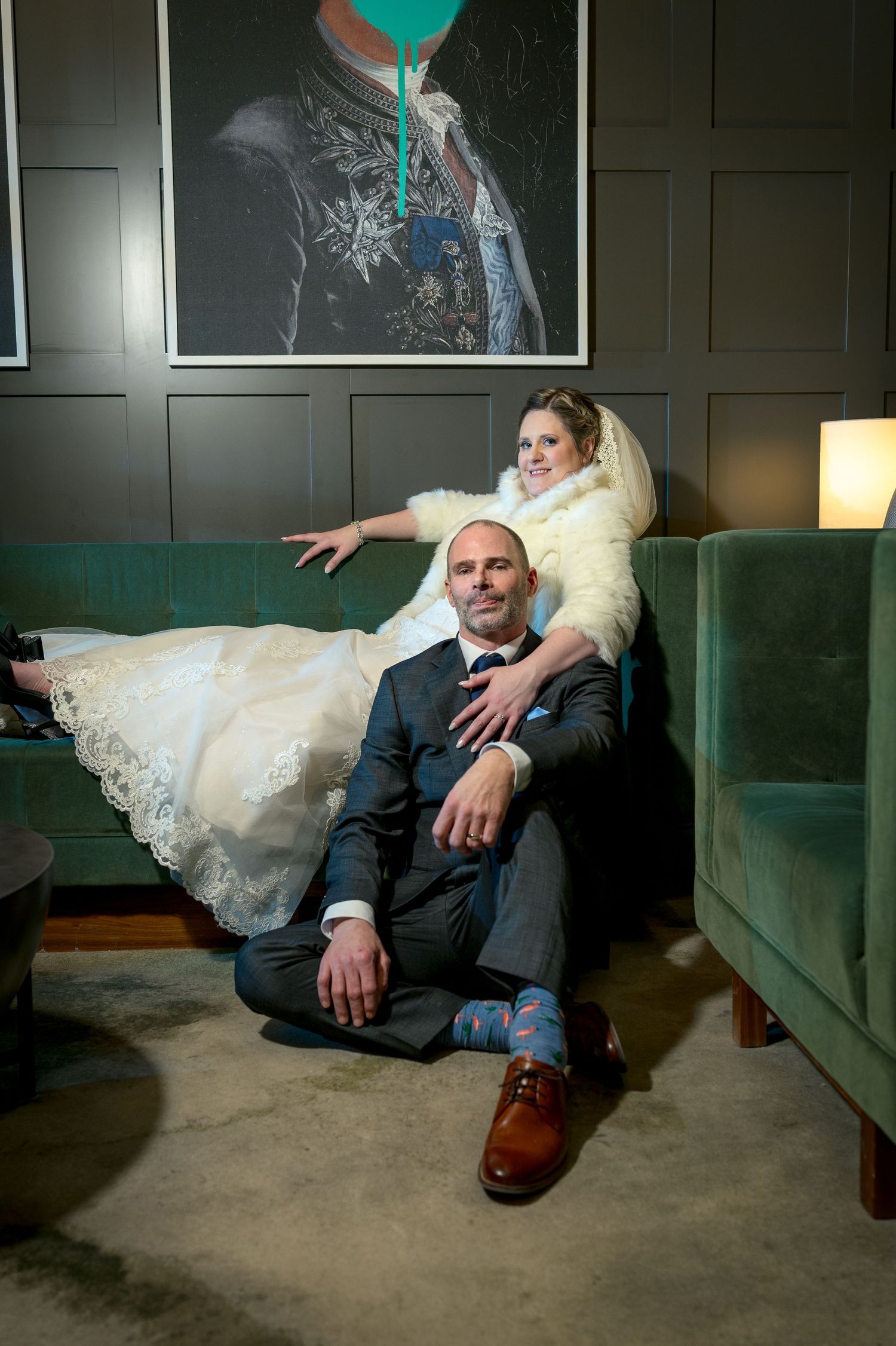 A couple in wedding attire relaxes casually on a green velvet couch in a dimly lit room with dark walls. Ottawa Wedding Photographer