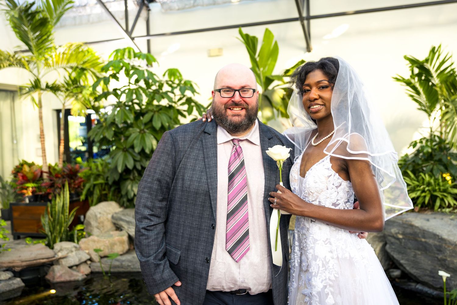 Bride & Groom pose together in formal attire at aquatopia wedding reception with lush greenery and windows in the background. Tim Gowling