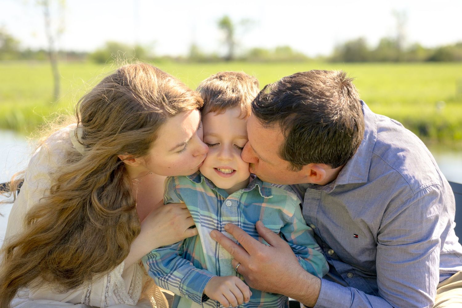 Parents kissing their young son on the cheeks outdoors with a green field in the background. Ottawa Family Photographer