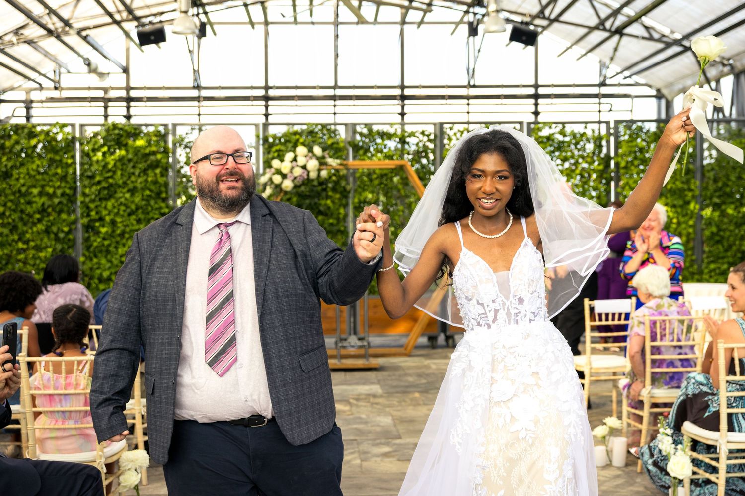 A celebratory moment captured in a greenhouse venue as newlyweds raise their hands in joy during their wedding ceremony.