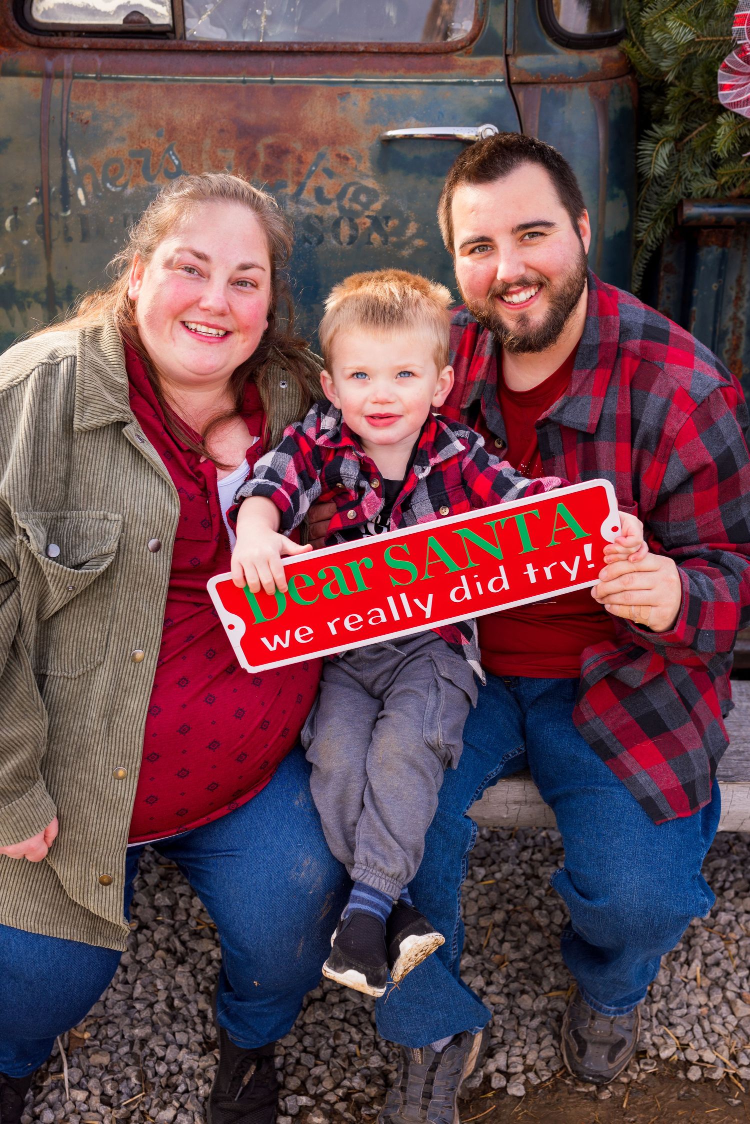 A family poses together outdoors with a red 'Dear Santa' sign by an old blue truck during a Christmas photo session.