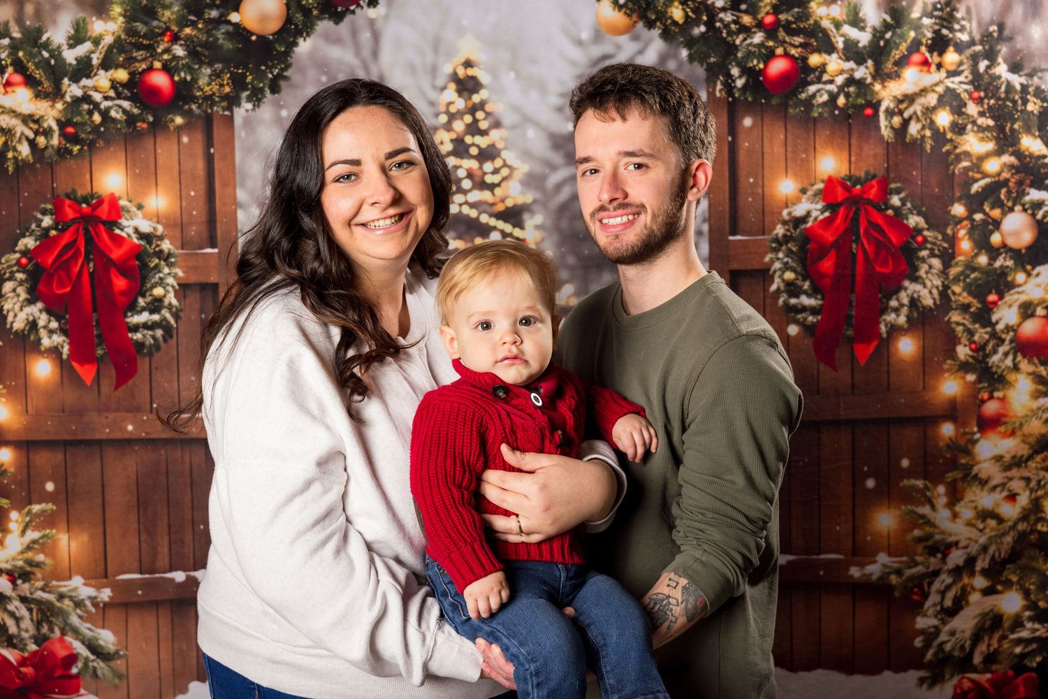 Family poses for portrait in front of rustic wood doors decorated with Christmas garland, red bows and holiday lights.