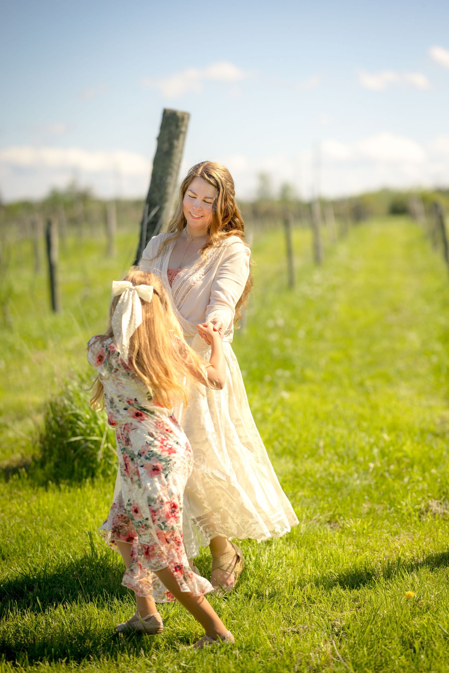 A mother and young daughter in floral dresses dancing joyfully in a sunny green vineyard on a bright summer day. Ottawa Family Photographer