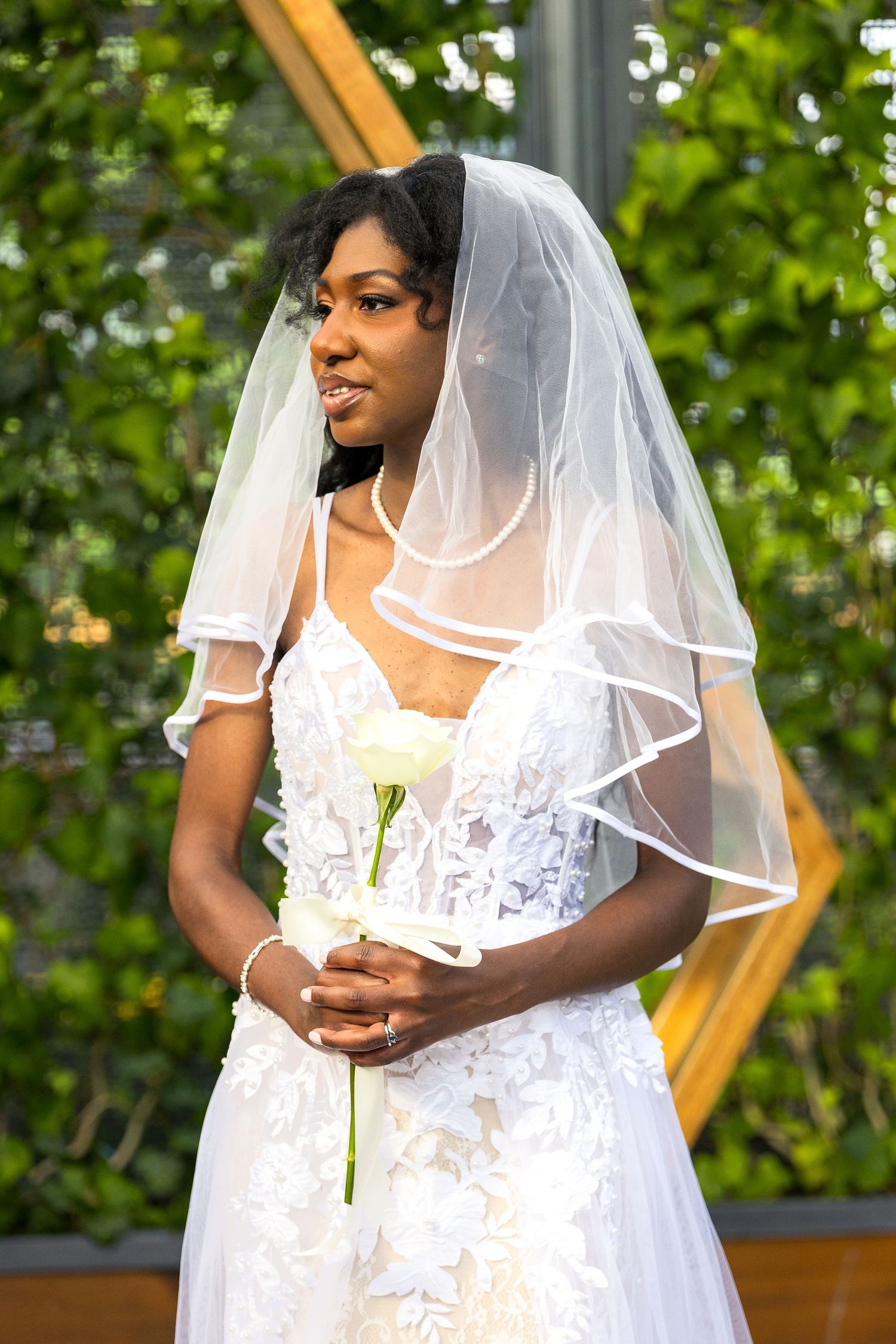 A bride in a white lace wedding dress and sheer veil at the altar in Aquatopia wedding venue with green foliage in the background.