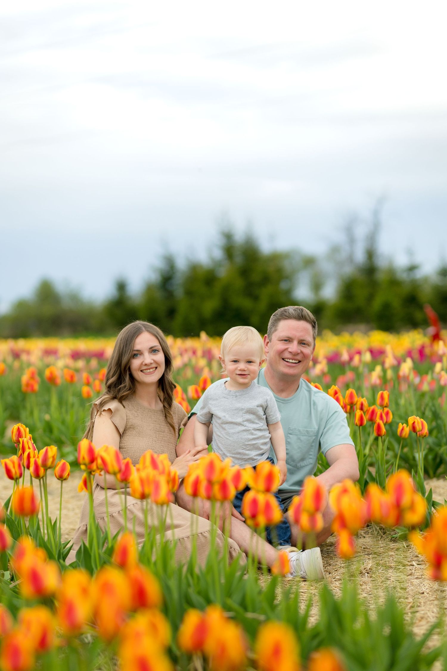 Family relaxing together between rows of vibrant orange tulips in a spring flower field on a sunny day. Ottawa Portrait Photographer