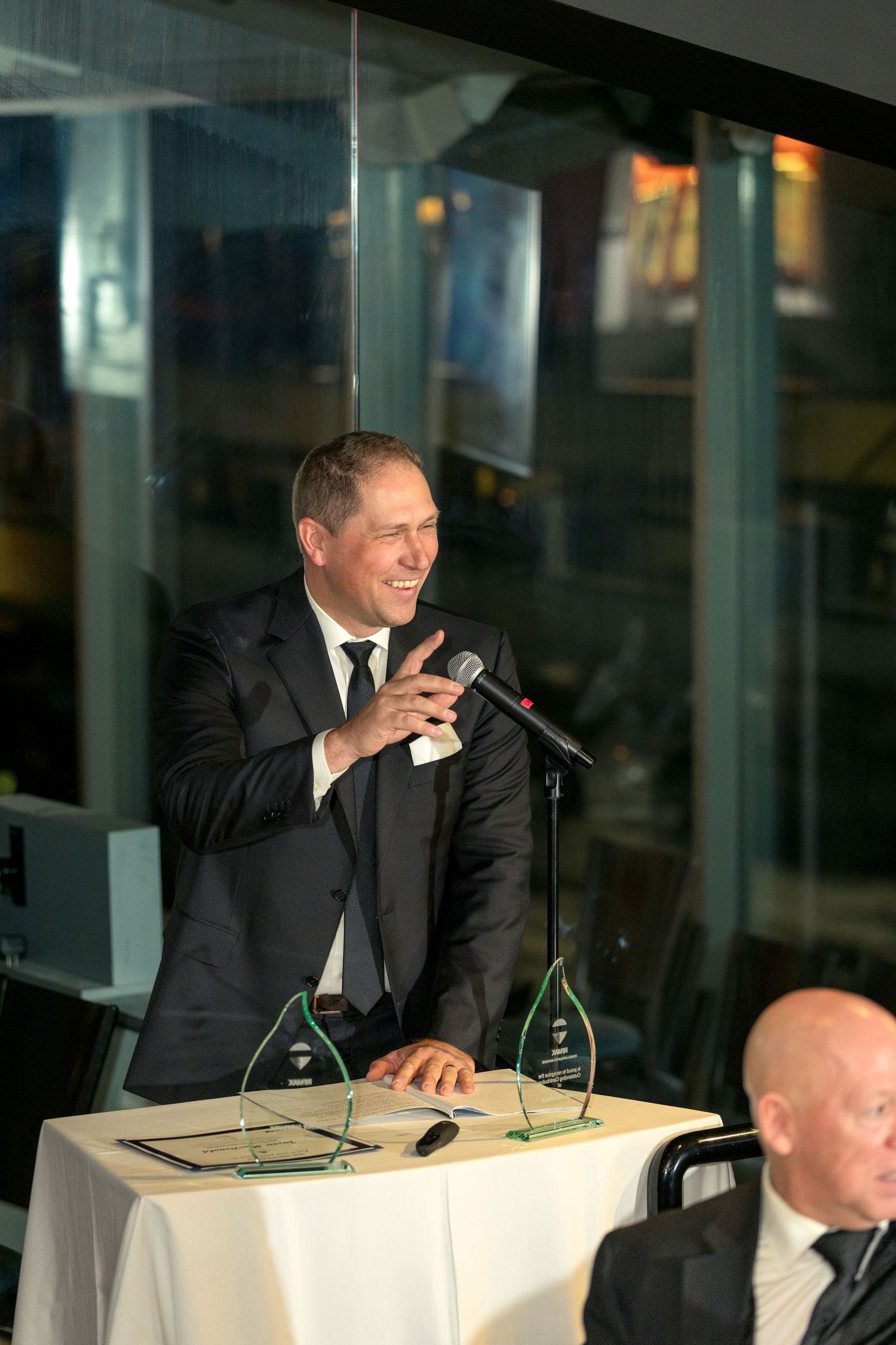 A speaker in a black suit delivers remarks at a podium with award trophies during an evening event. Ottawa event Photographer