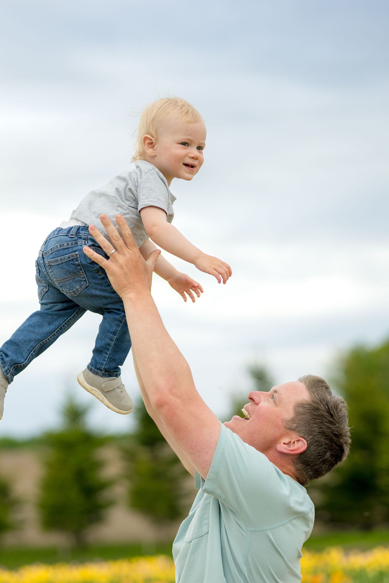 Parent playfully lifts baby up in the air while standing in a field of yellow tulips on a cloudy day. Ottawa portrait photographer