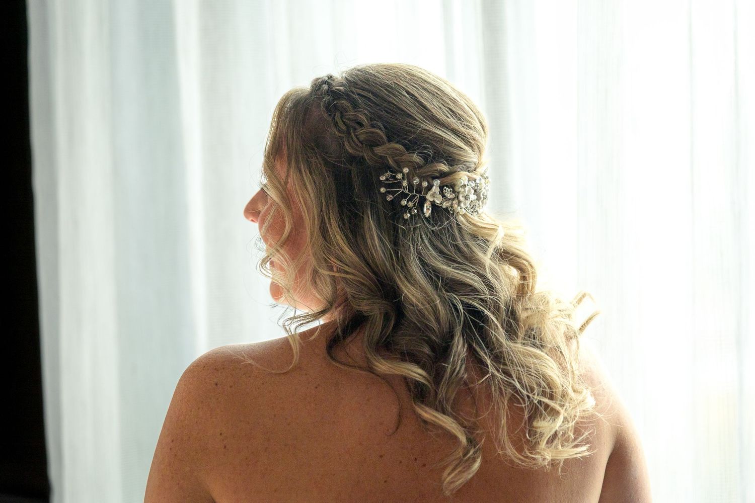 A bride with wavy blonde hair and sparkly hair accessories looks away from camera in front of white curtains. Ottawa wedding photographer