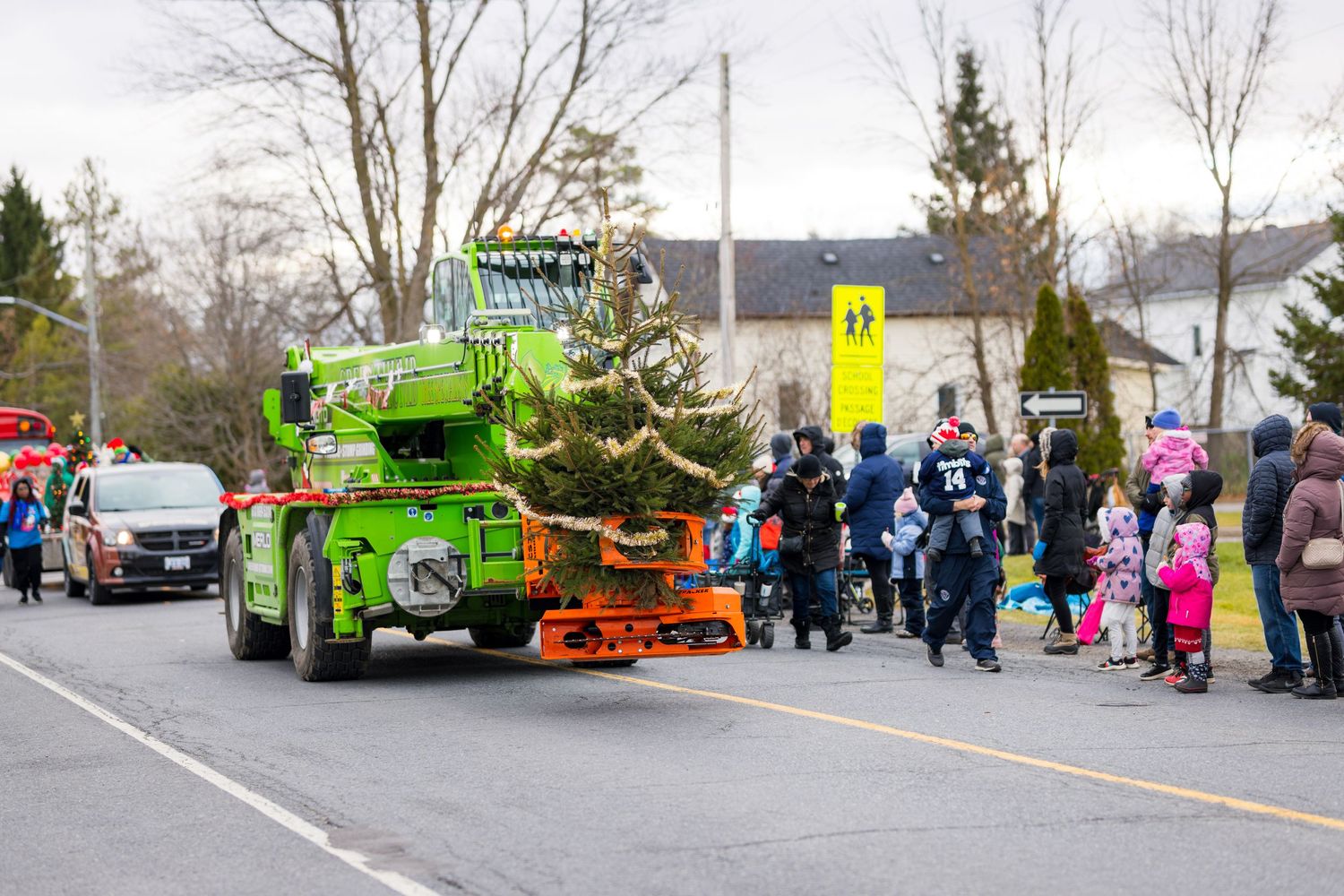 A bright green tow truck decorated as a festive Christmas tree drives past spectators at a holiday parade.
