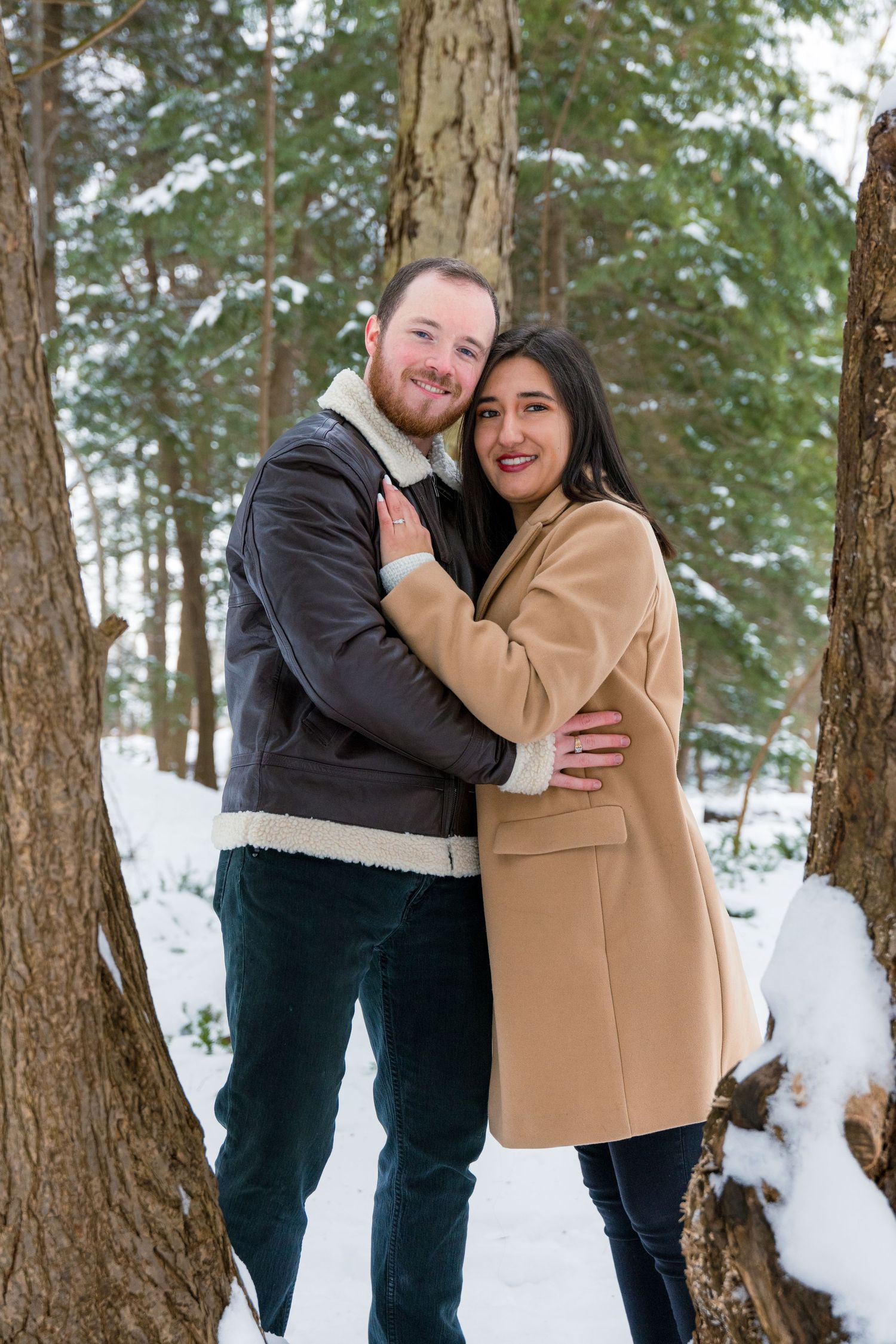 A couple embraces while standing between snowy trees in a winter forest setting.