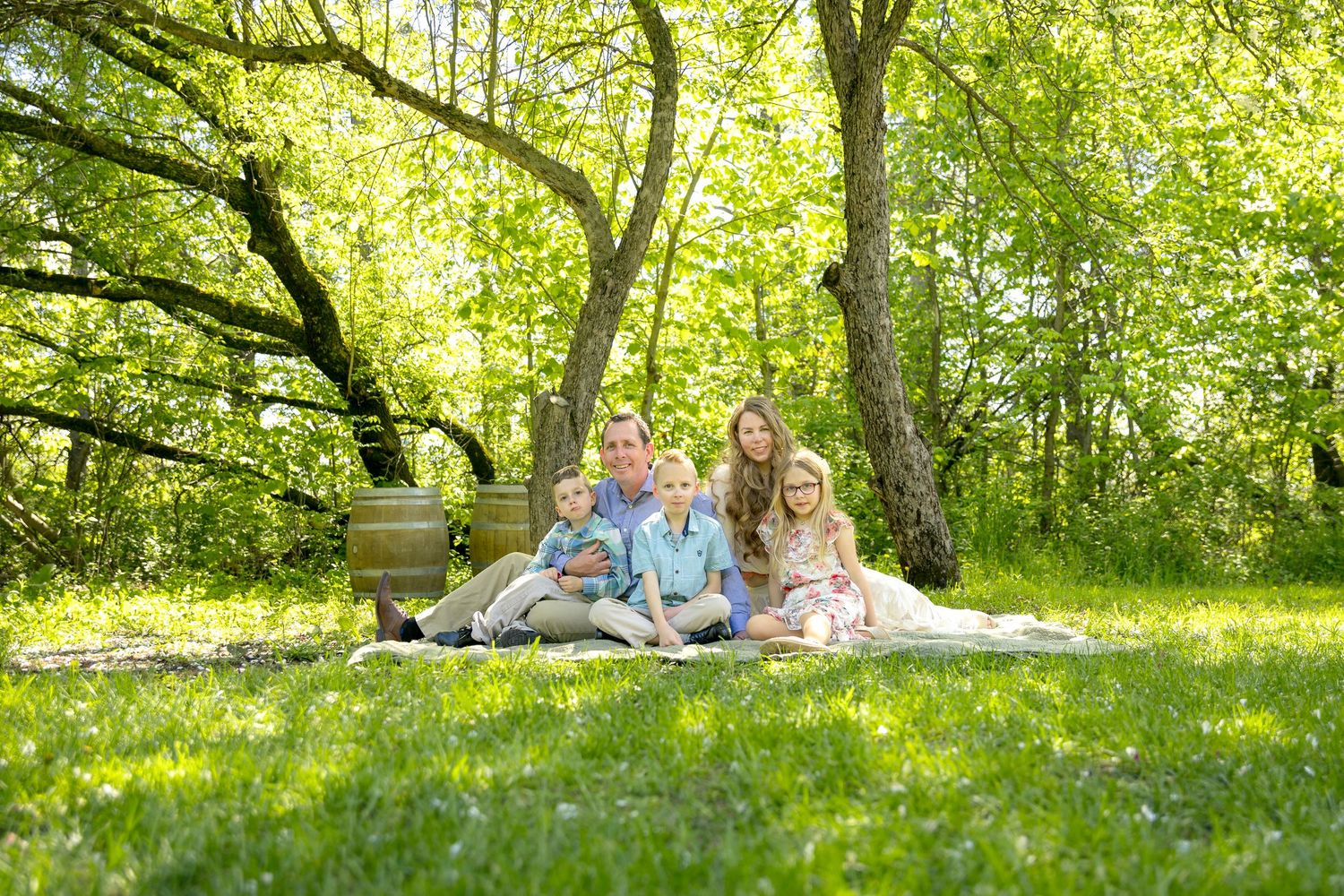Family of four smiling together on a blanket in a lush green park under leafy trees on a sunny day. Ottawa Family Photographer