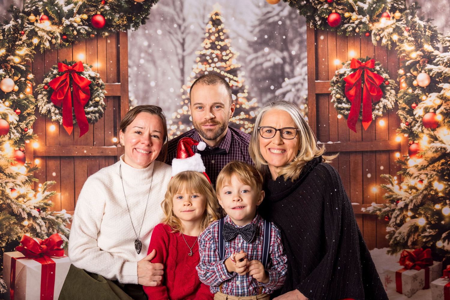 A family poses together for a holiday portrait in front of decorated doors with Christmas trees and red bows.