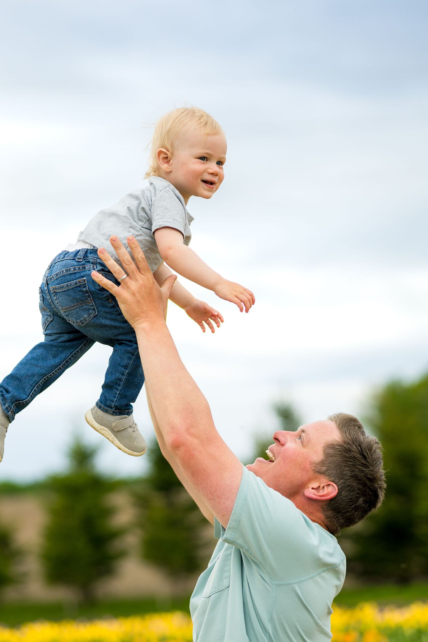 Playful outdoor moment as a parent lifts a small child high in the air against a backdrop of yellow wildflowers. Ottawa Photographer