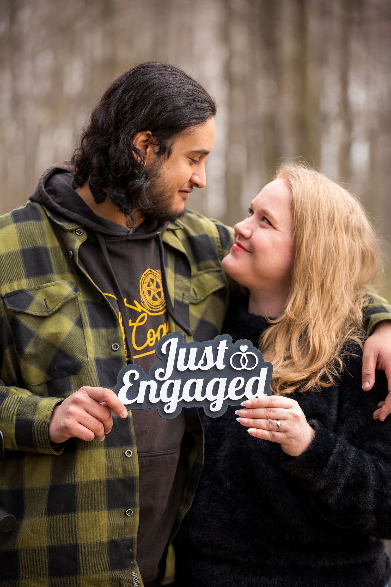 A couple in casual winter wear hold up a 'Just Engaged' sign while sharing a tender moment in the woods.