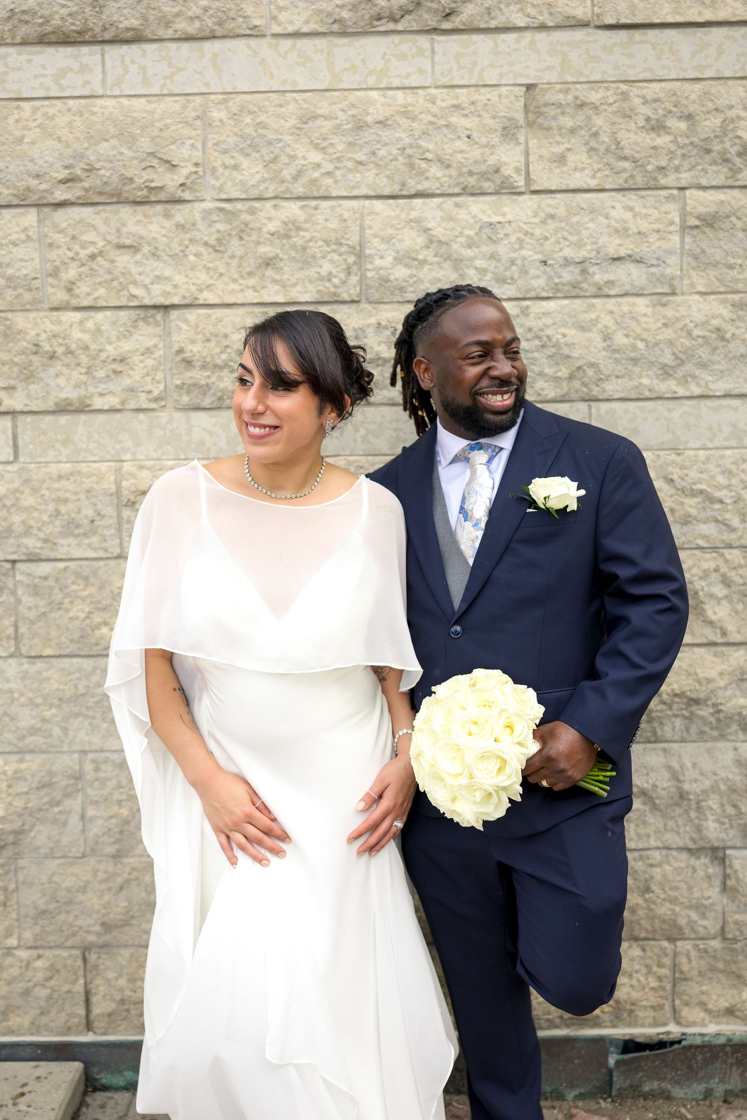 Happy interracial newlyweds pose against stone wall, bride in white cape dress holding bouquet, groom in navy suit. Ottawa Wedding Photo