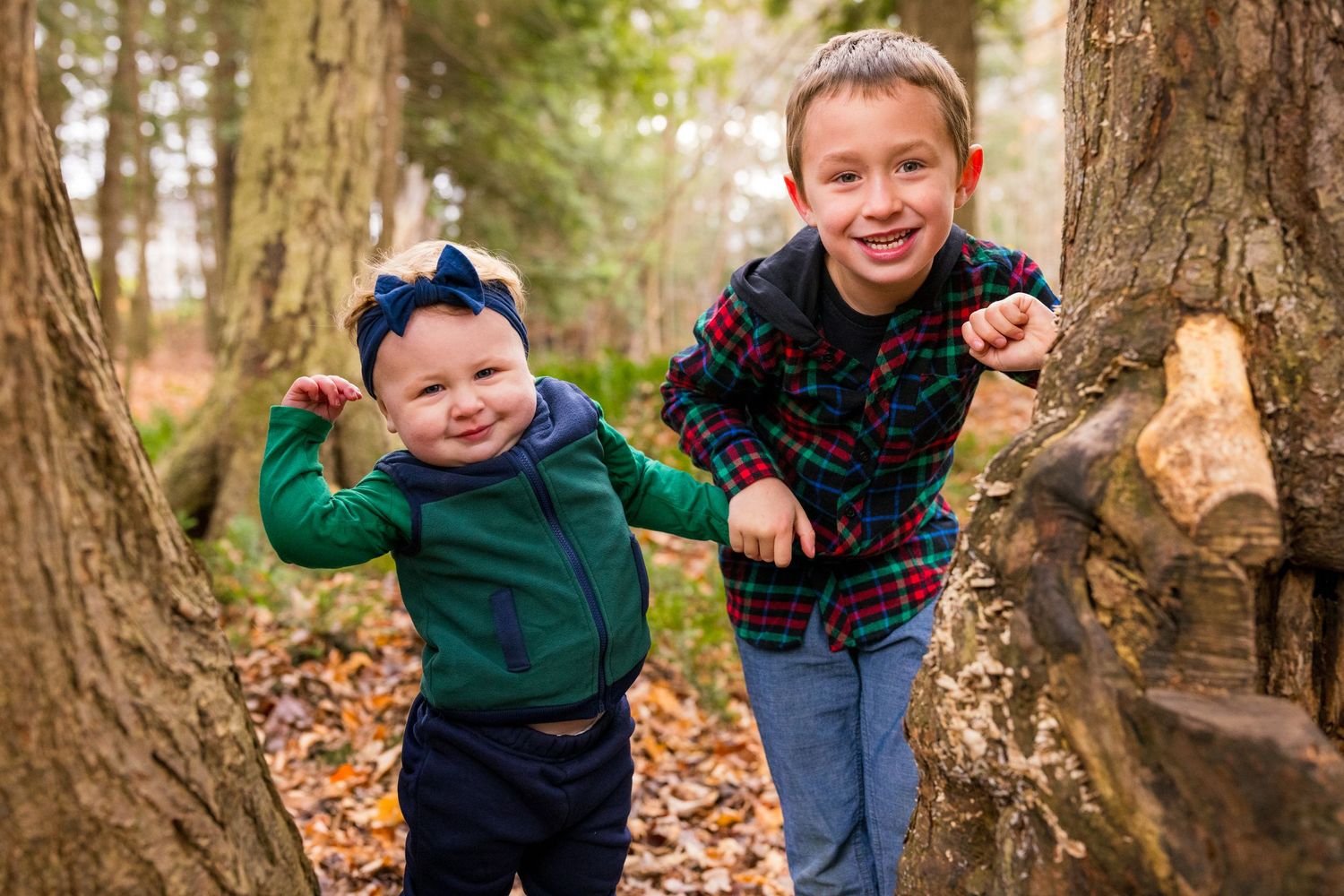 Two young children play and laugh between large tree trunks in a forest covered with autumn leaves.