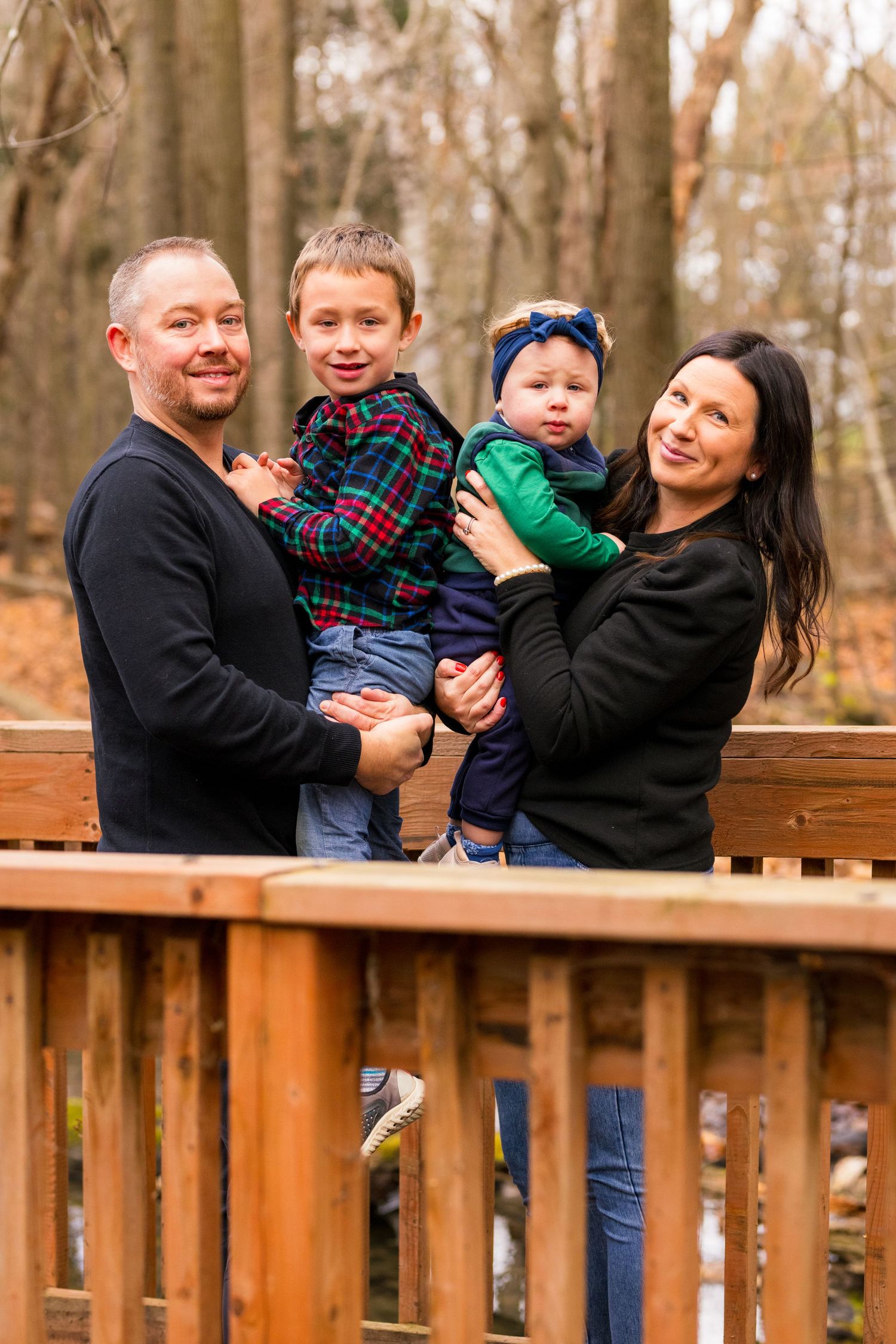 A family stands together smiling on a wooden deck in an autumn forest setting during a photo shoot.