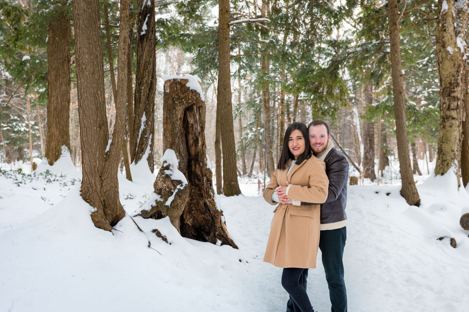 A couple embraces on a snowy winter trail surrounded by tall trees in a serene forest landscape.