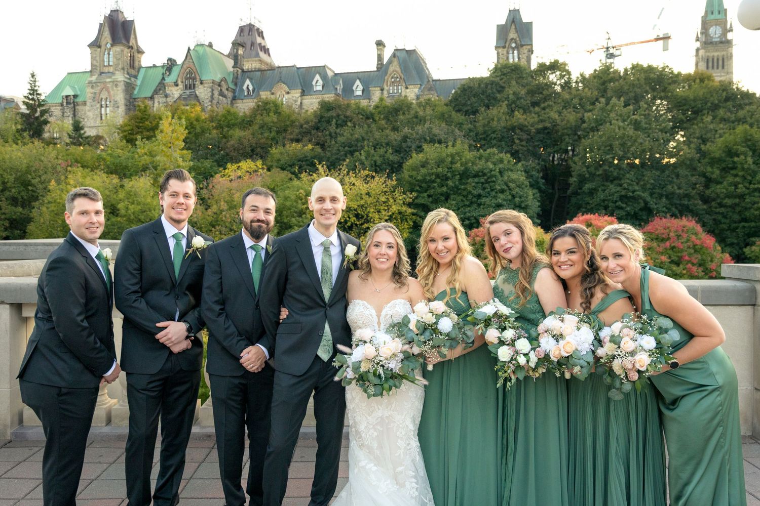 A wedding party in sage green dresses and black suits poses together outdoors on a cloudy day. Ottawa Wedding Photographer