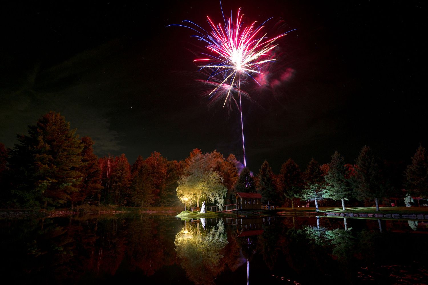 Colorful fireworks burst over a reflective lake at Bean Town Receptions, illuminating autumn trees with vibrant red and pink hues.