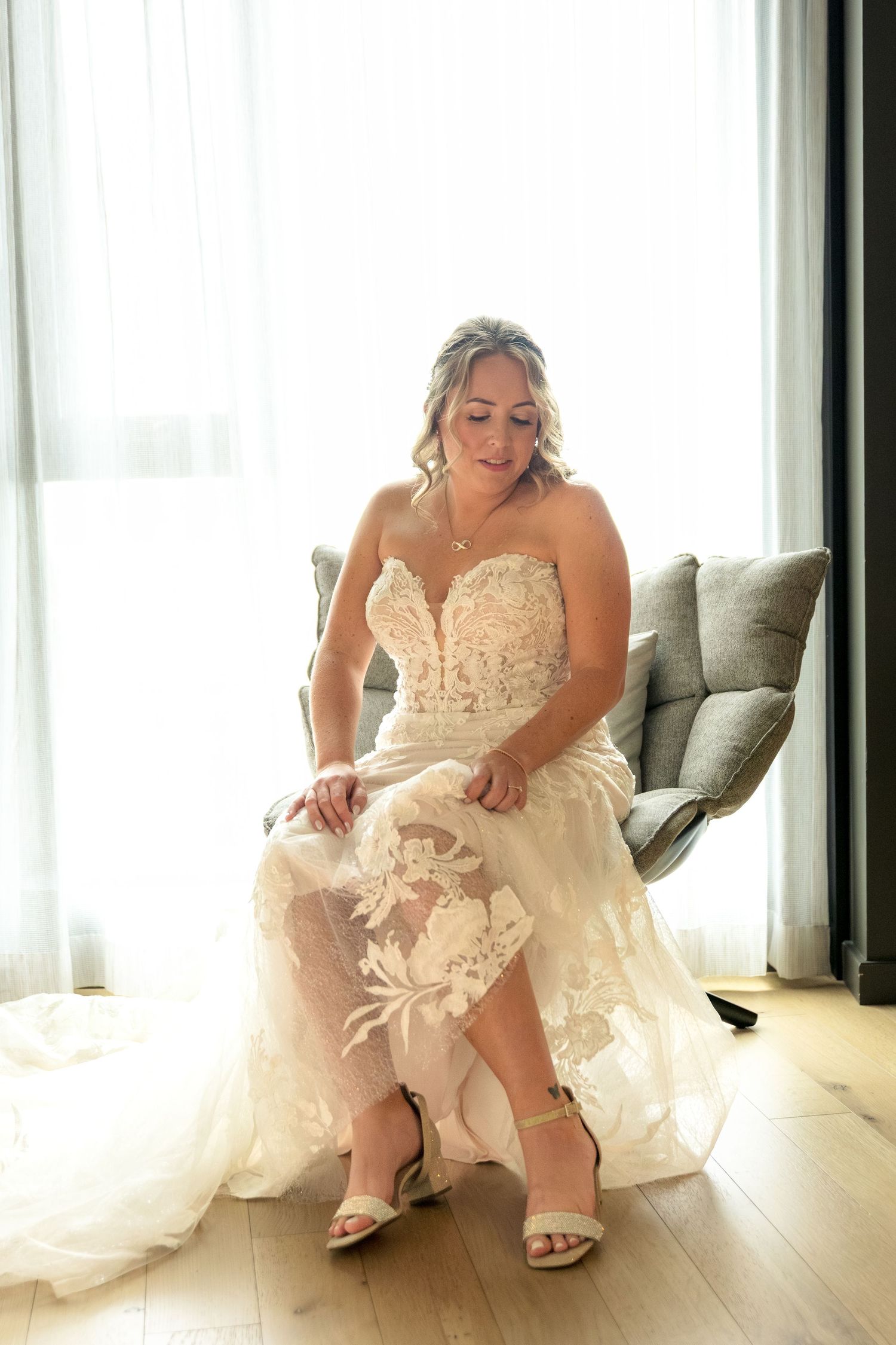 A bride in a white lace gown sits on a couch while putting on elegant heels in a bright, modern room. Ottawa Wedding Photographer