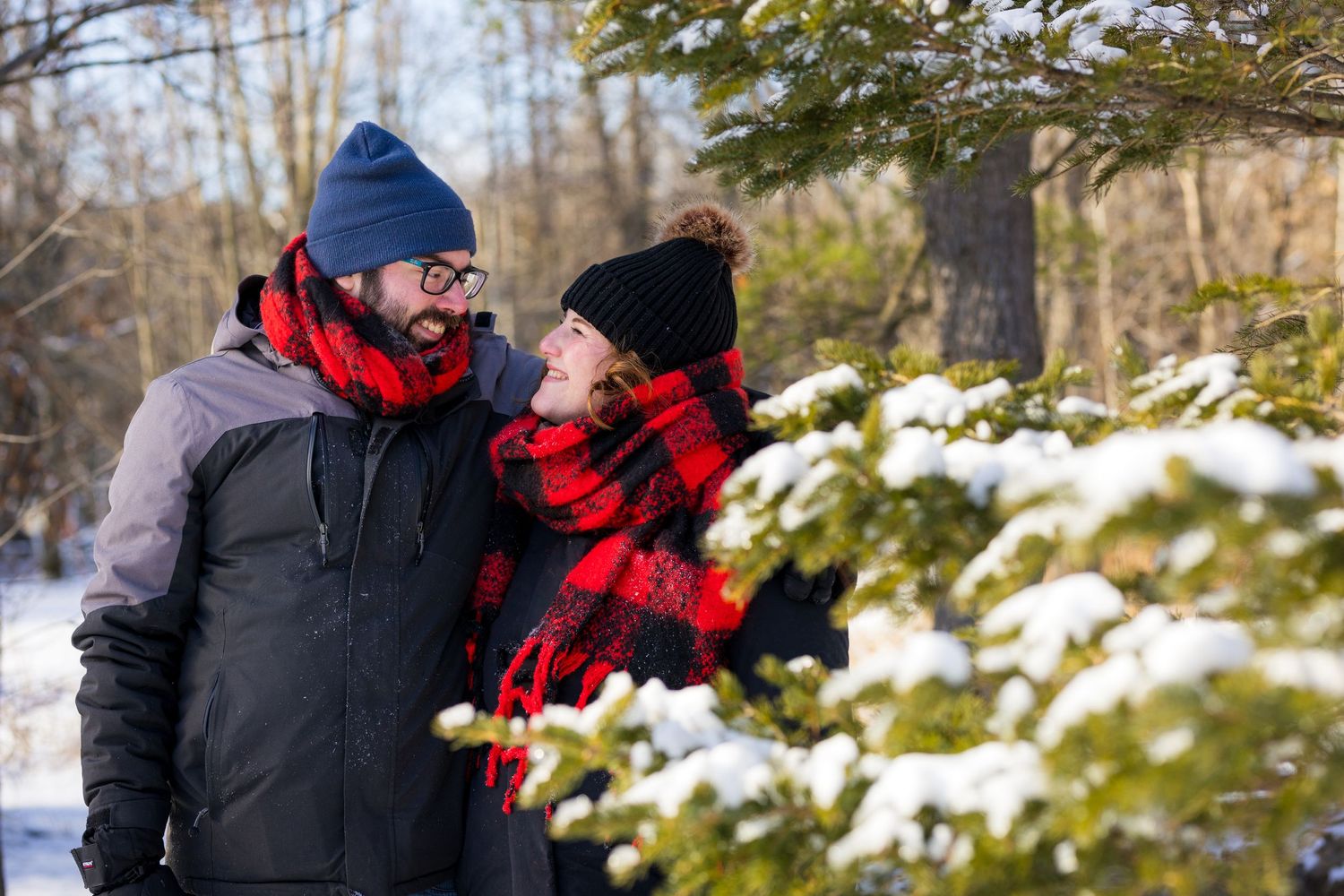 Two people in winter coats and hats stand together in a snowy landscape beside snow-covered evergreen branches.
