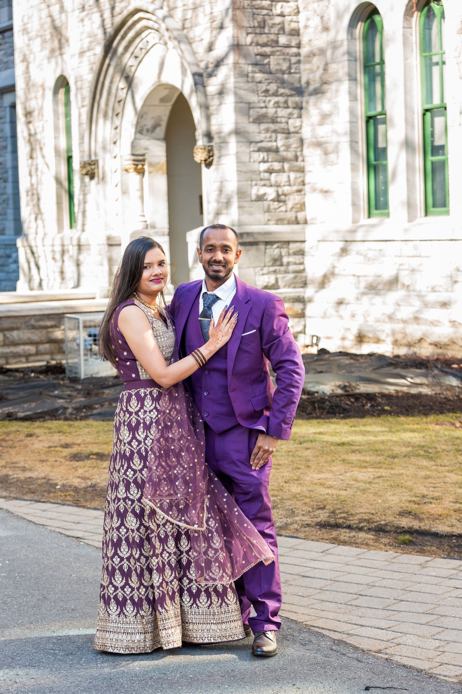 A couple poses outdoors in formal attire with purple suit and patterned burgundy dress.