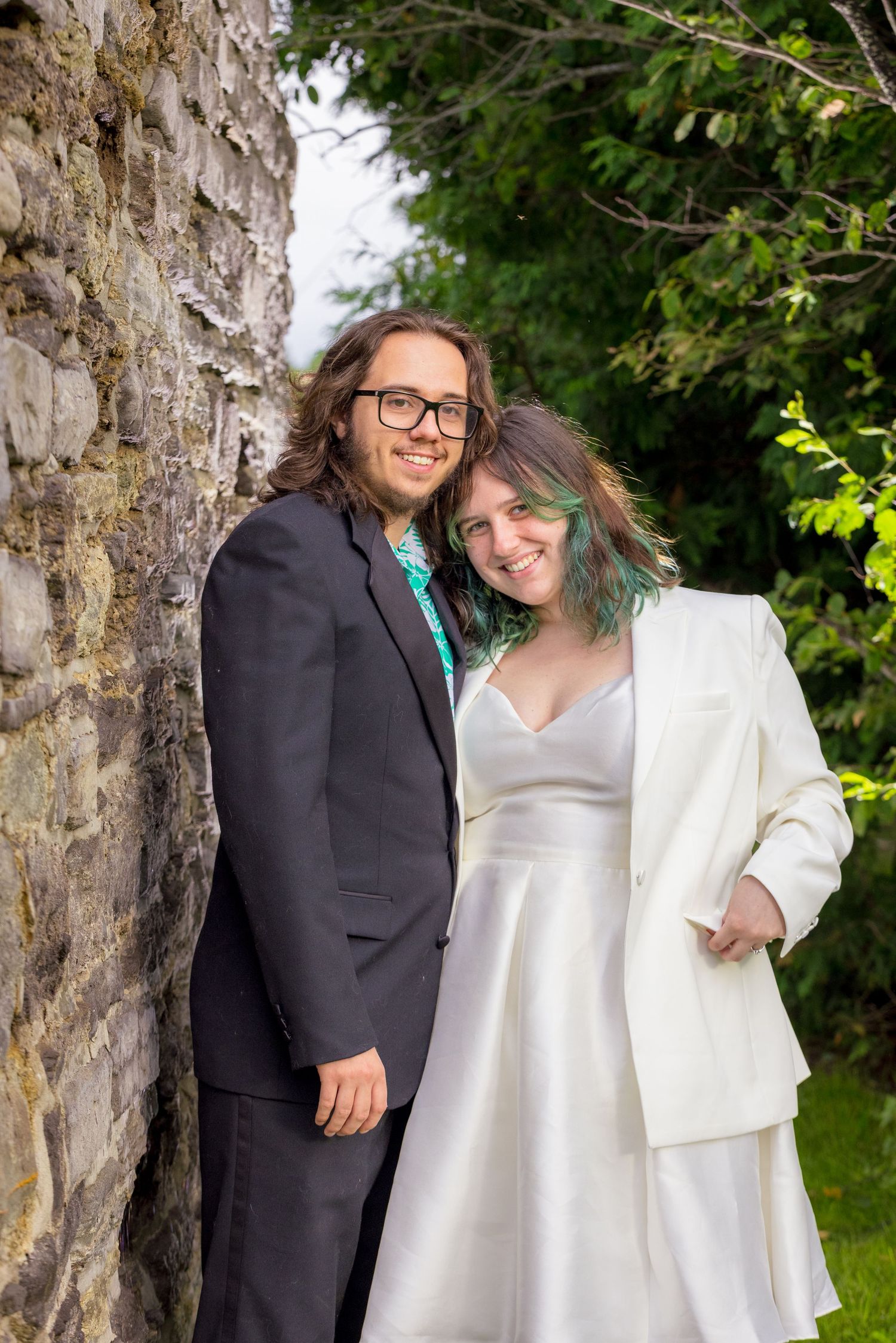 A couple in wedding attire poses against a stone wall in a garden setting. Ottawa Wedding Photographer