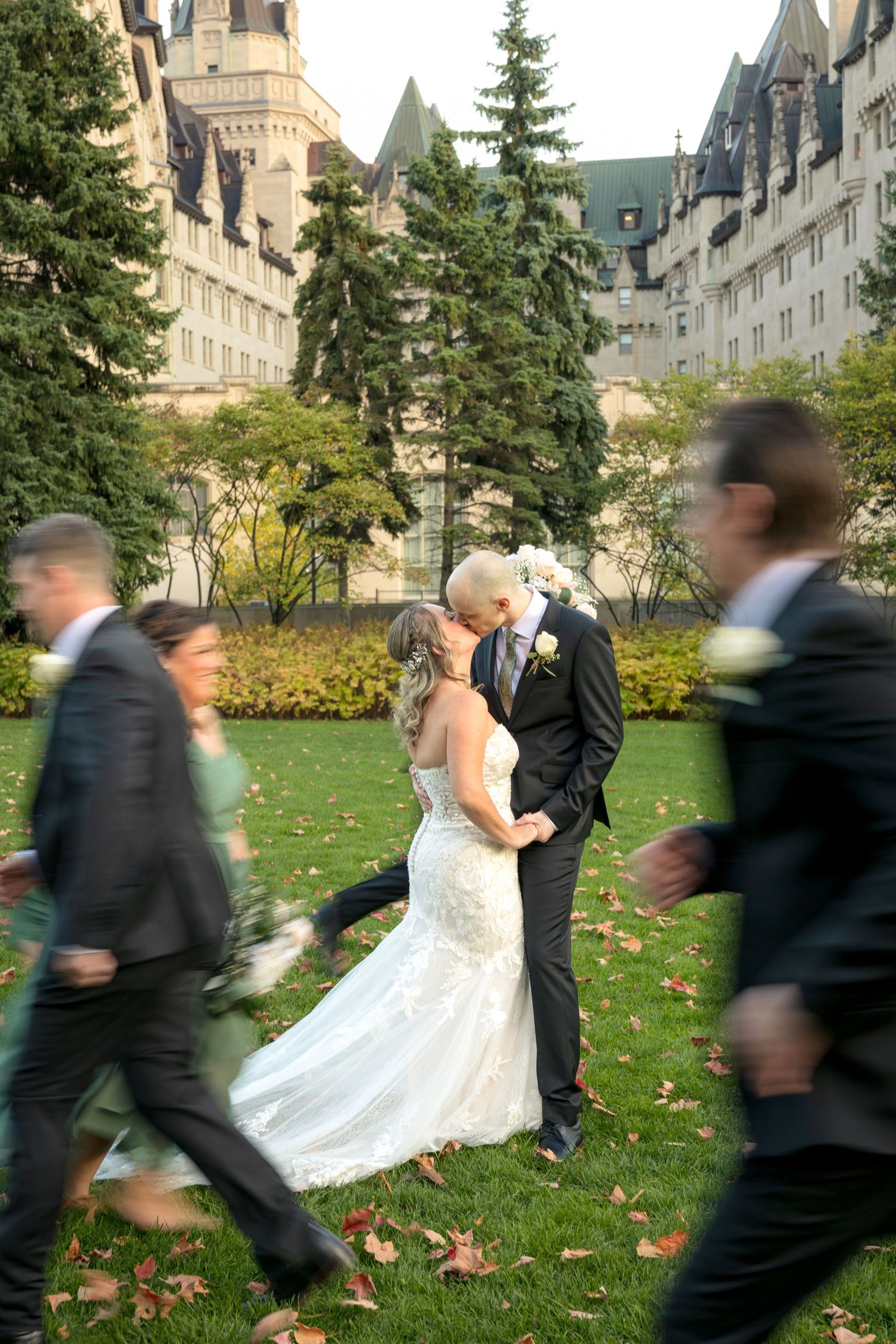 Wedding couple embraces on autumn lawn while guests walk past in motion blur behind Fairmont Château Laurier Ottawa wedding photographer