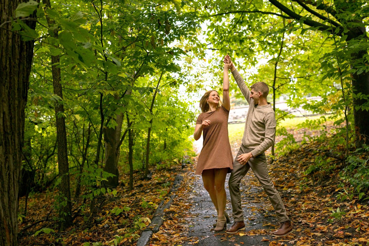 A playful couple dances on a leaf-covered forest trail surrounded by green trees. Ottawa Wedding Photographer
