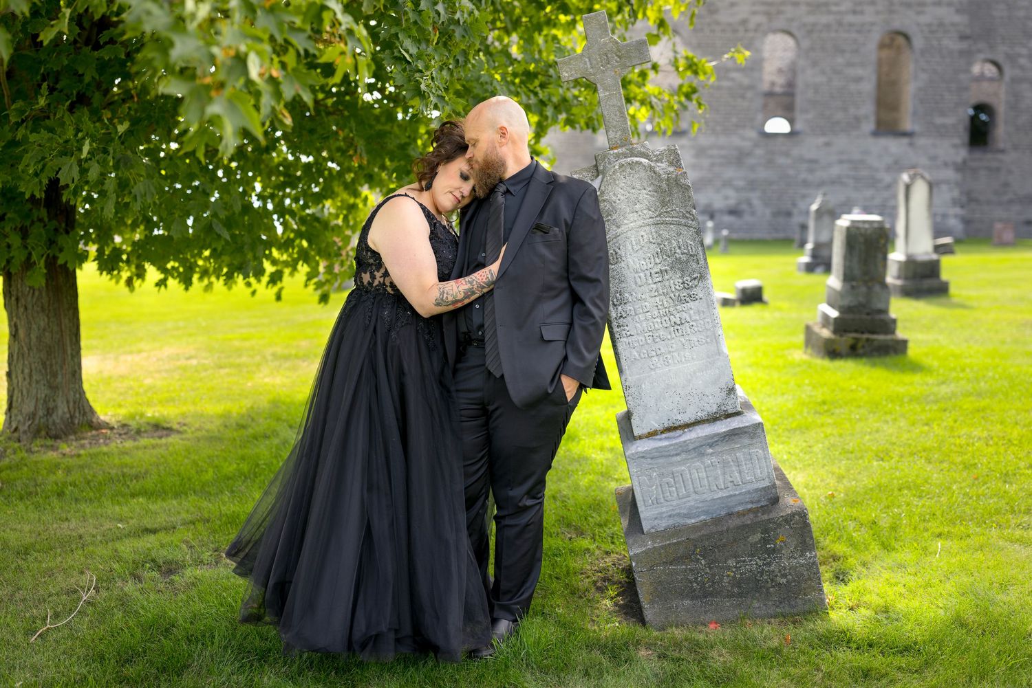 A couple in dark clothing walks through a cemetery with green grass and gray headstones on a sunny day. Ottawa Photographer Tim Gowling